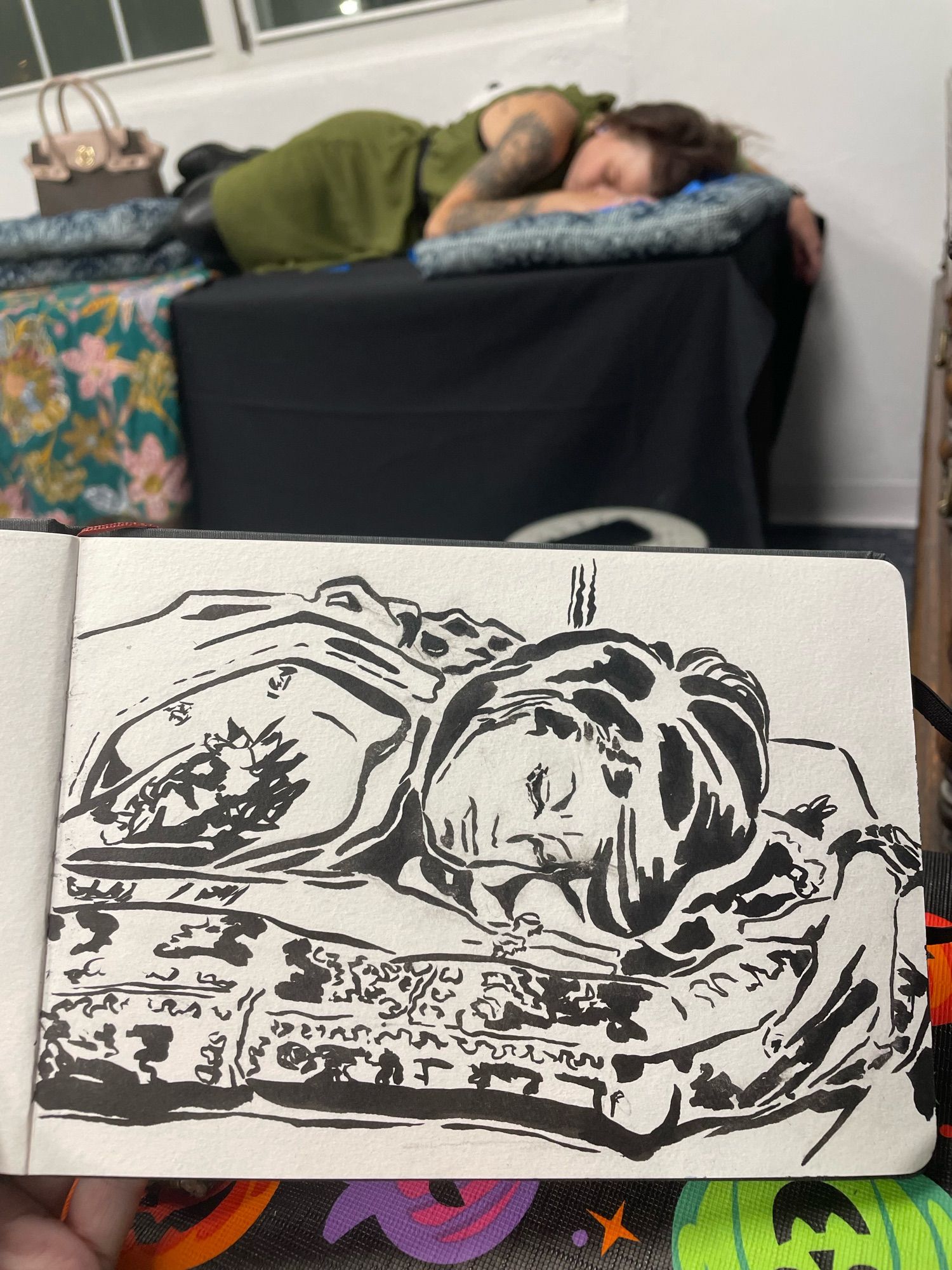 Ink drawing of a tattooed woman reclining on a table. She rests on a patterned pillow. Her hair is straight and short. Her left arm wraps around the pillow and wears a watch. The woman being drawn is posing behind the held sketch.