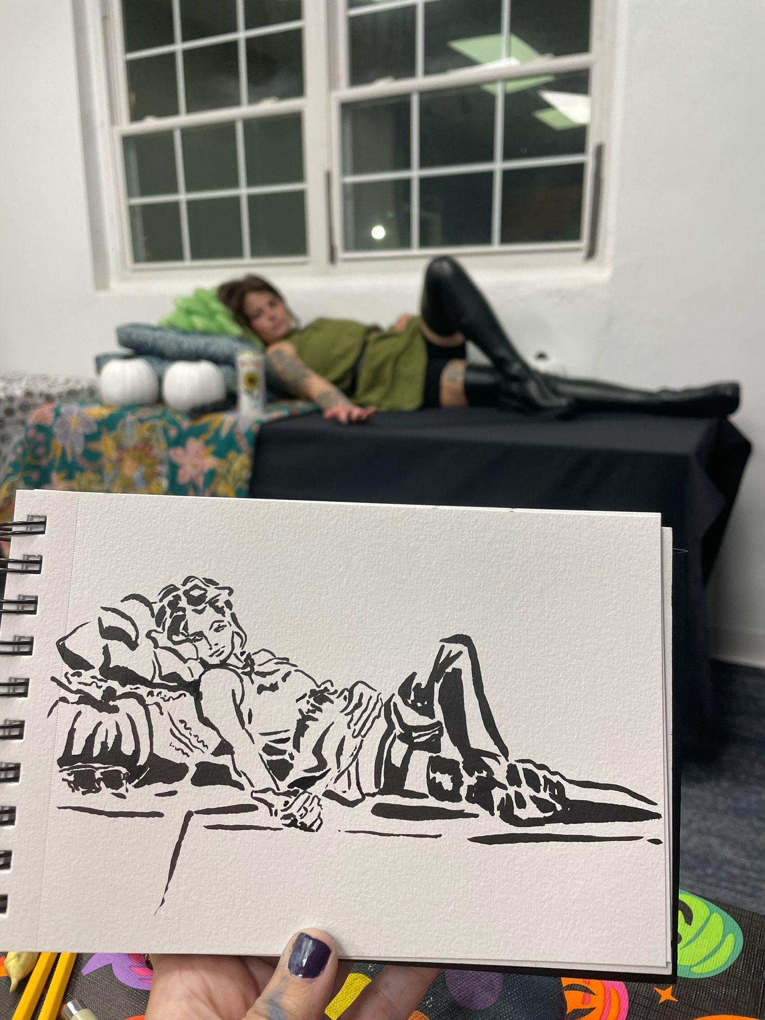 Ink drawing of a reclining woman on a table with a bunch of blankets and pillows. She is in thigh high boots, black shorts, and a green dress. The sketch is held in front of the posing model.