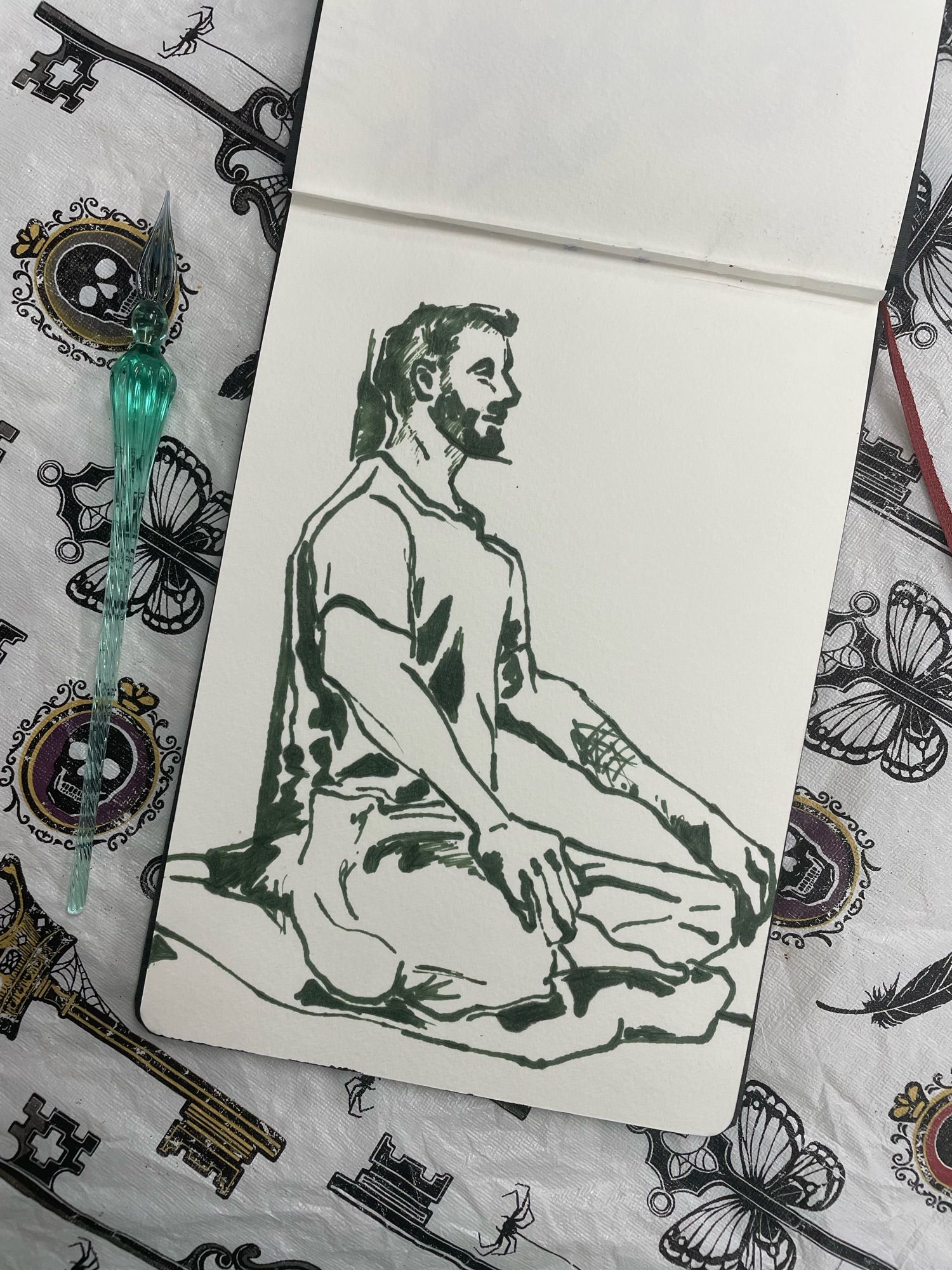 Green ink drawing of a man sitting cross legged with back straight against the wall. He has a geometric tattoo on his left forearm. The pen used to draw the figure is on its left.