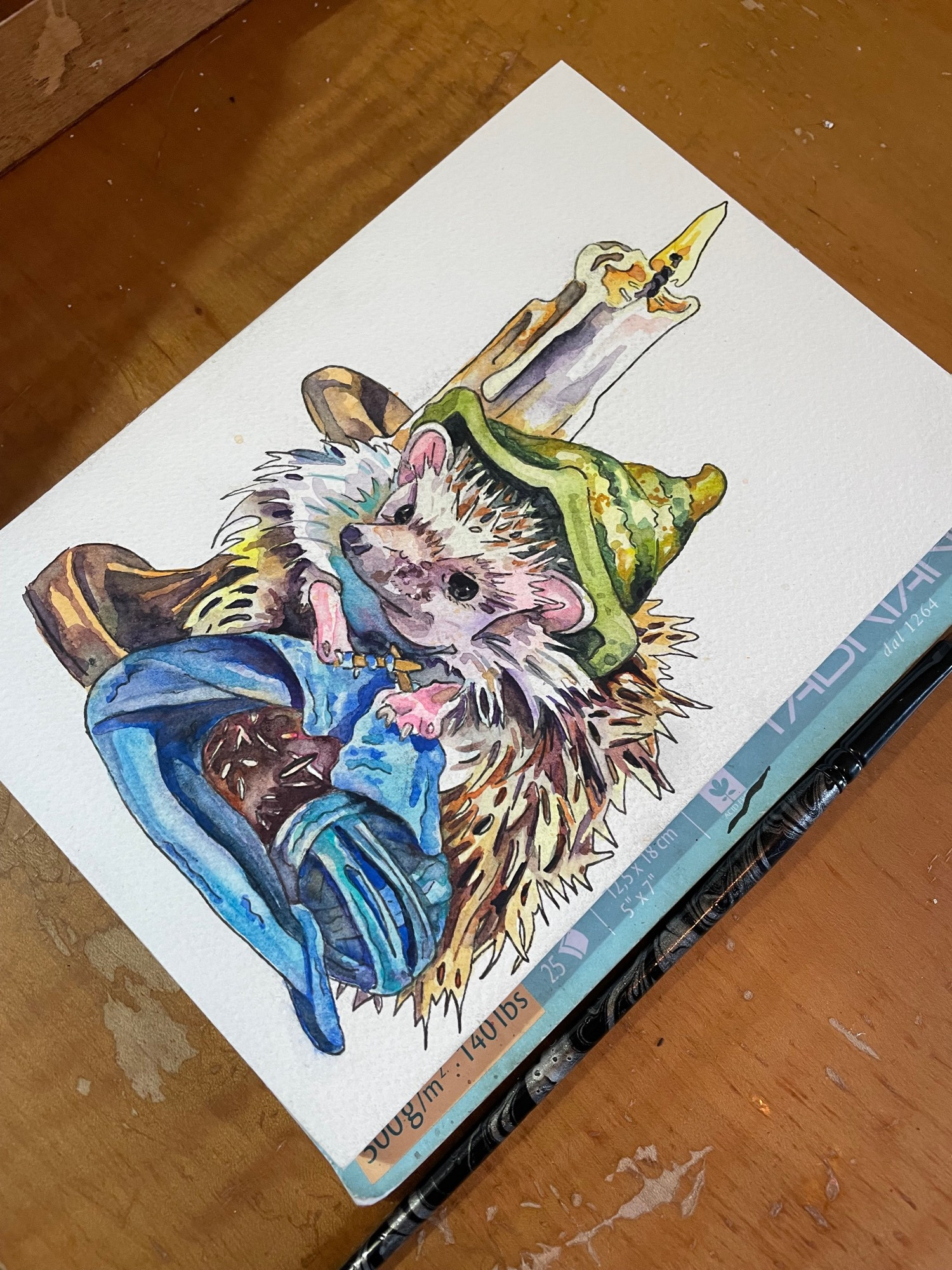 Unfinished watercolor and ink painting next to a paint brush. The illustration of a hedgehog wearing a witch’s hat knitting a blue scarf. A candle in a brass holder shines behind the animal.