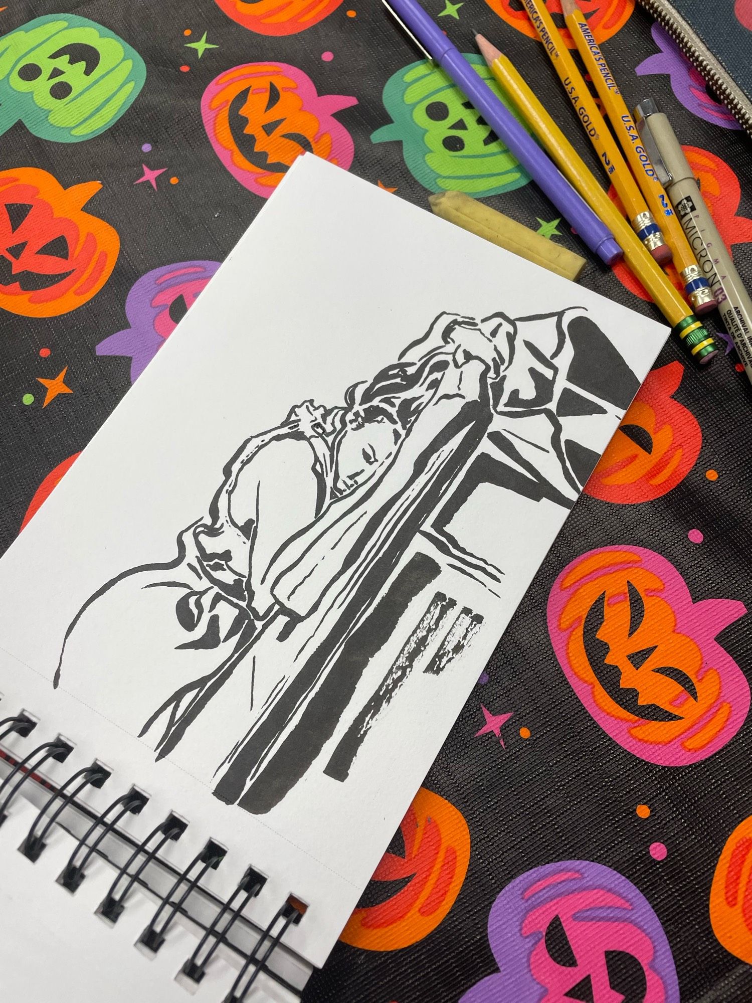 Ink drawing of a reclining woman asleep on a table. It is in a sketchbook on a table decorated with jack o lanterns next to pens and pencils.