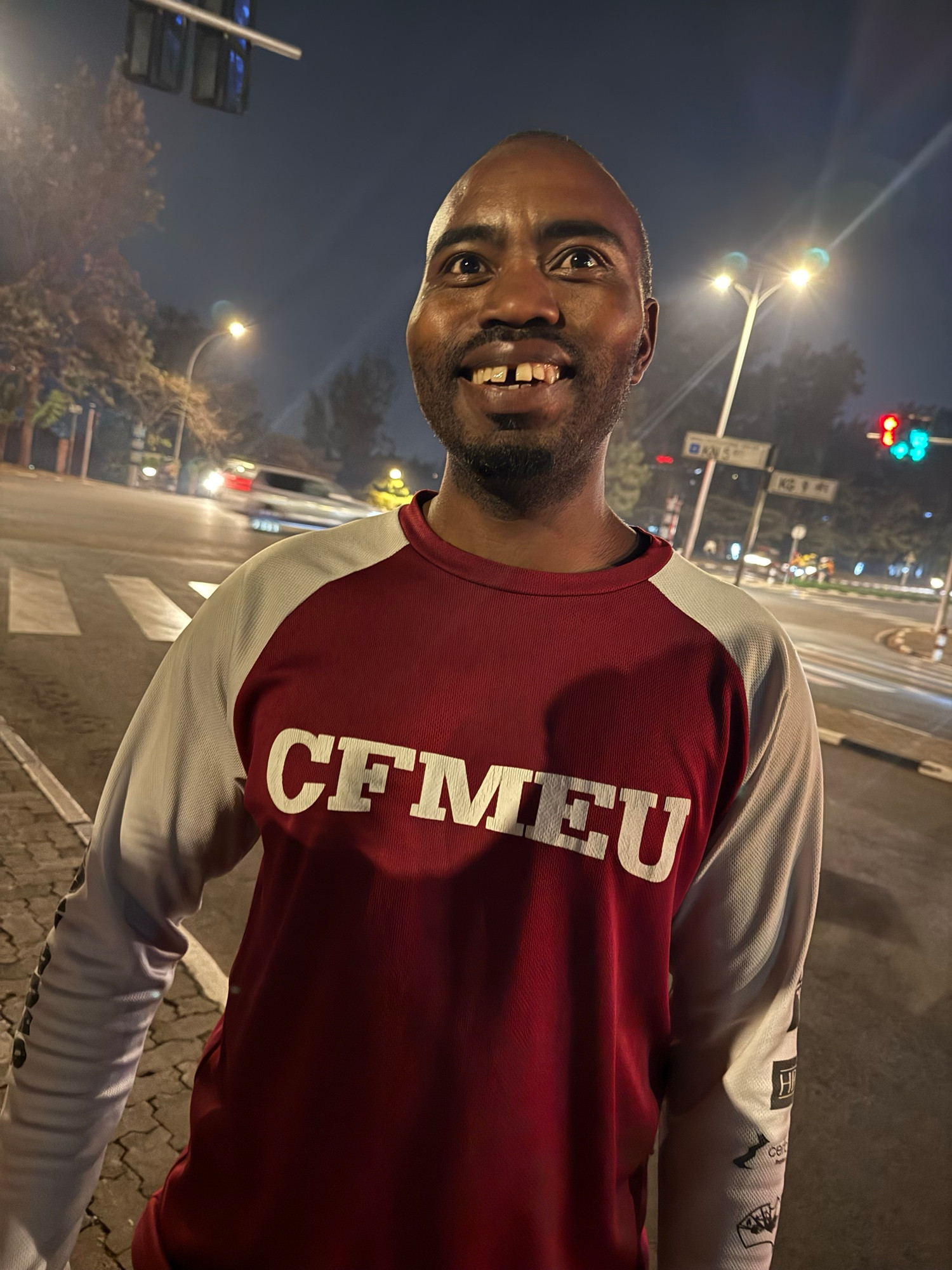 Kigali, Rwanda. A man stands on a city street at night, smiling warmly at the camera. He is wearing a maroon and white long-sleeve shirt with the letters “CFMEU” printed in bold white font across the chest. The background features a well-lit urban intersection with traffic lights, cars, and trees visible under the glow of streetlights. The atmosphere is lively and illuminated by the artificial lights against a dark sky.