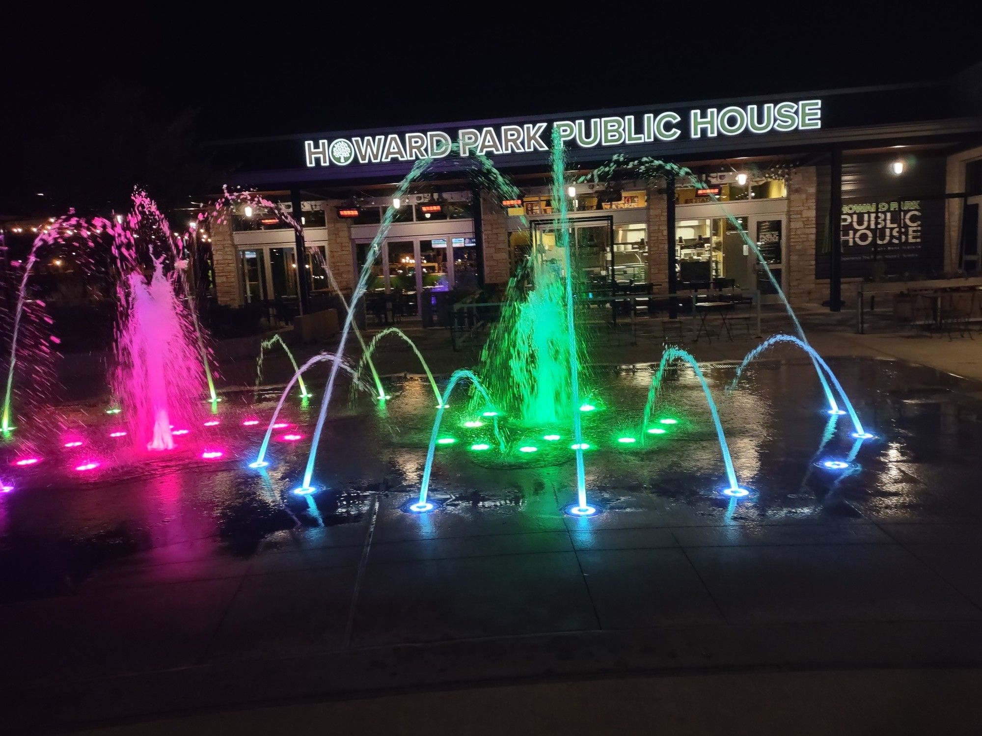 Fountains and lights splashing red, blue, and green water around with a restaurant called "Howard Park Public House" in the background