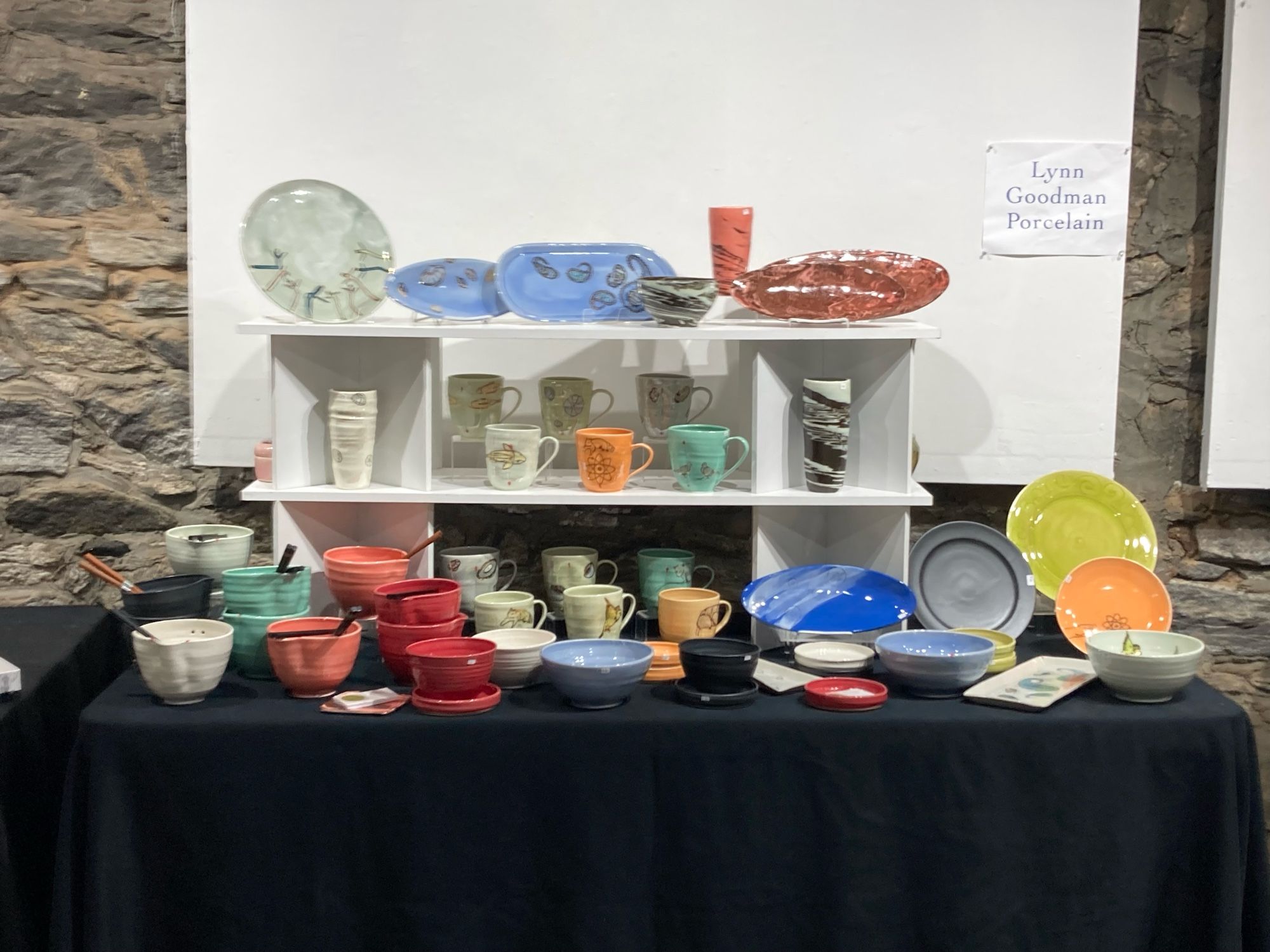 Packed display of assorted colorful handmade pottery; table with black tablecloth, then two white shelves above it, covered with pots.