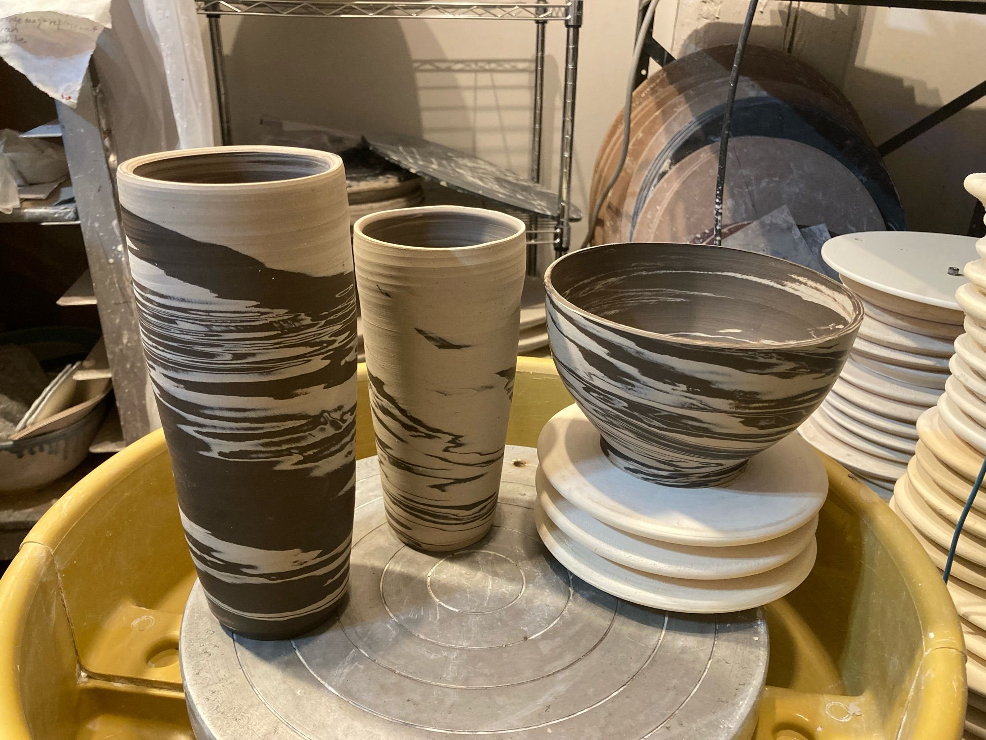 Three pieces of thrown and trimmed marbleized clay, on top of a potter’s wheel. The clay is dark brown and porcelain white. Background is pottery tools and storage.