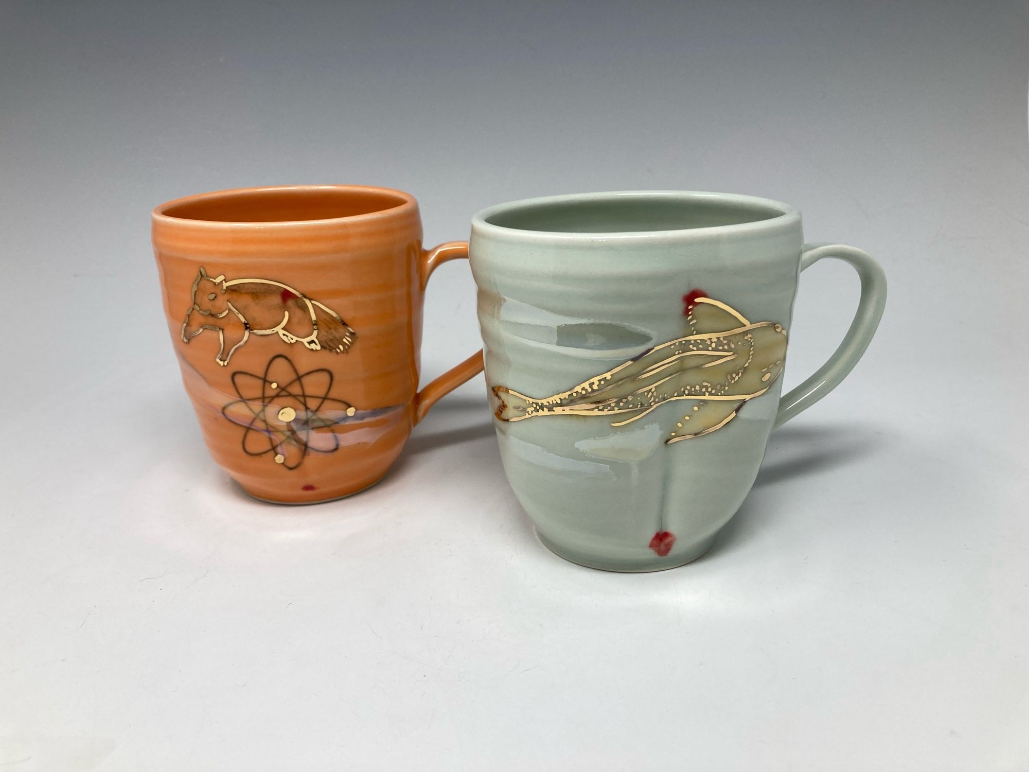 Two thrown porcelain mugs. On the left, “atomic squirrel”, in orange, which has a squirrel and an atom diagram, red dots, with gold highlights. On the right “koi”, in celadon,  with red dots, with gold highlights