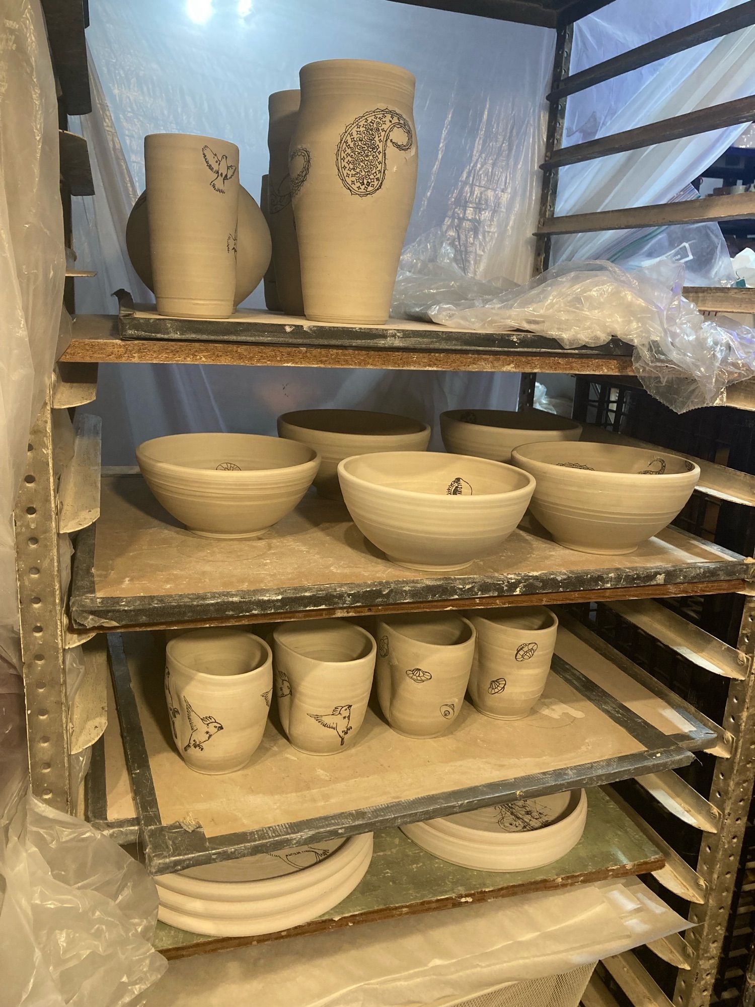 A ware cart, full of unfired pottery. Top: vases. Next shelf: bowls. Next shelf: mugs. Final shelf: stacked platters.