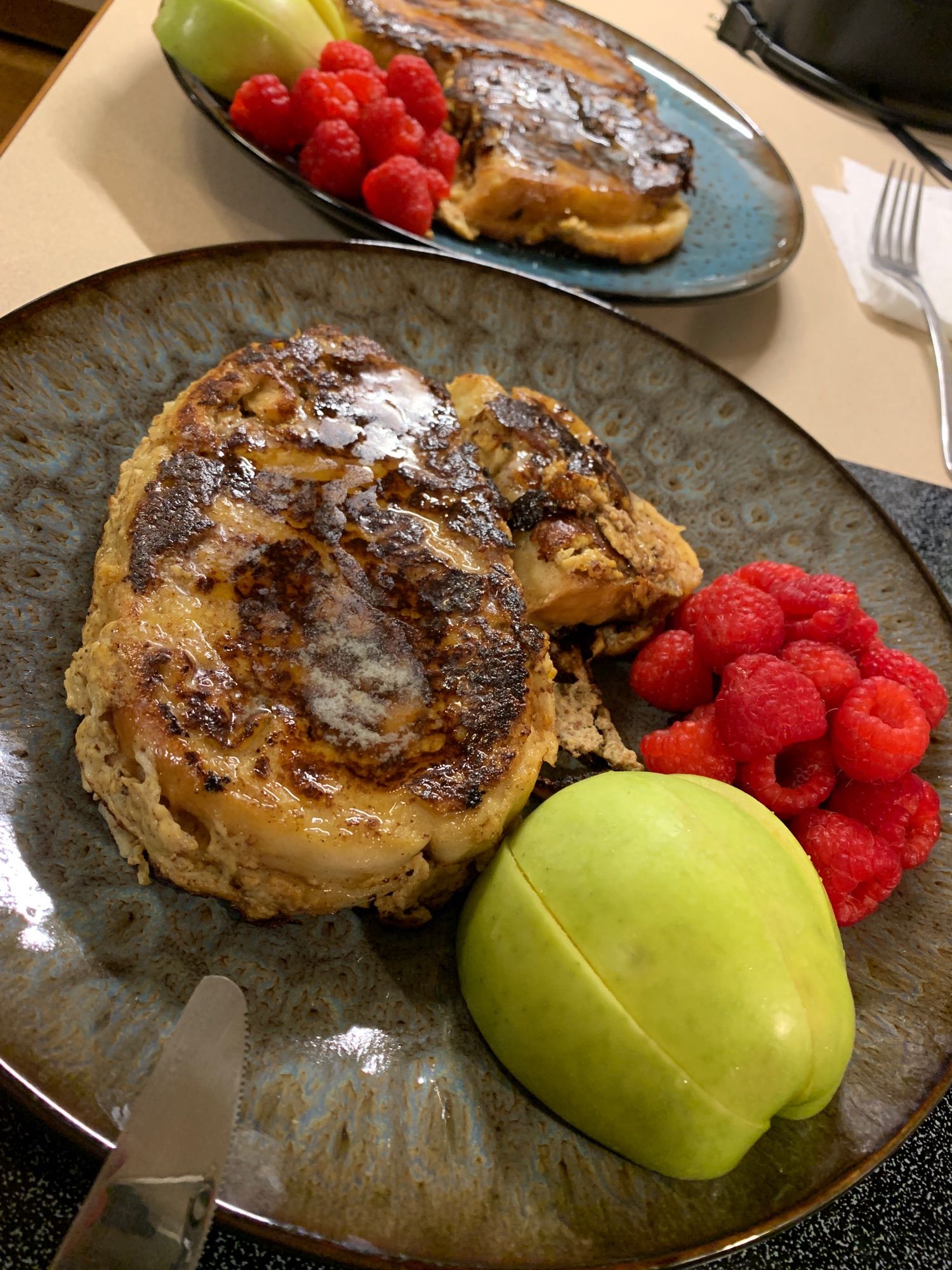 Two plates of French toast topped with glistening butter and syrup, with raspberries and green apple slices on the side