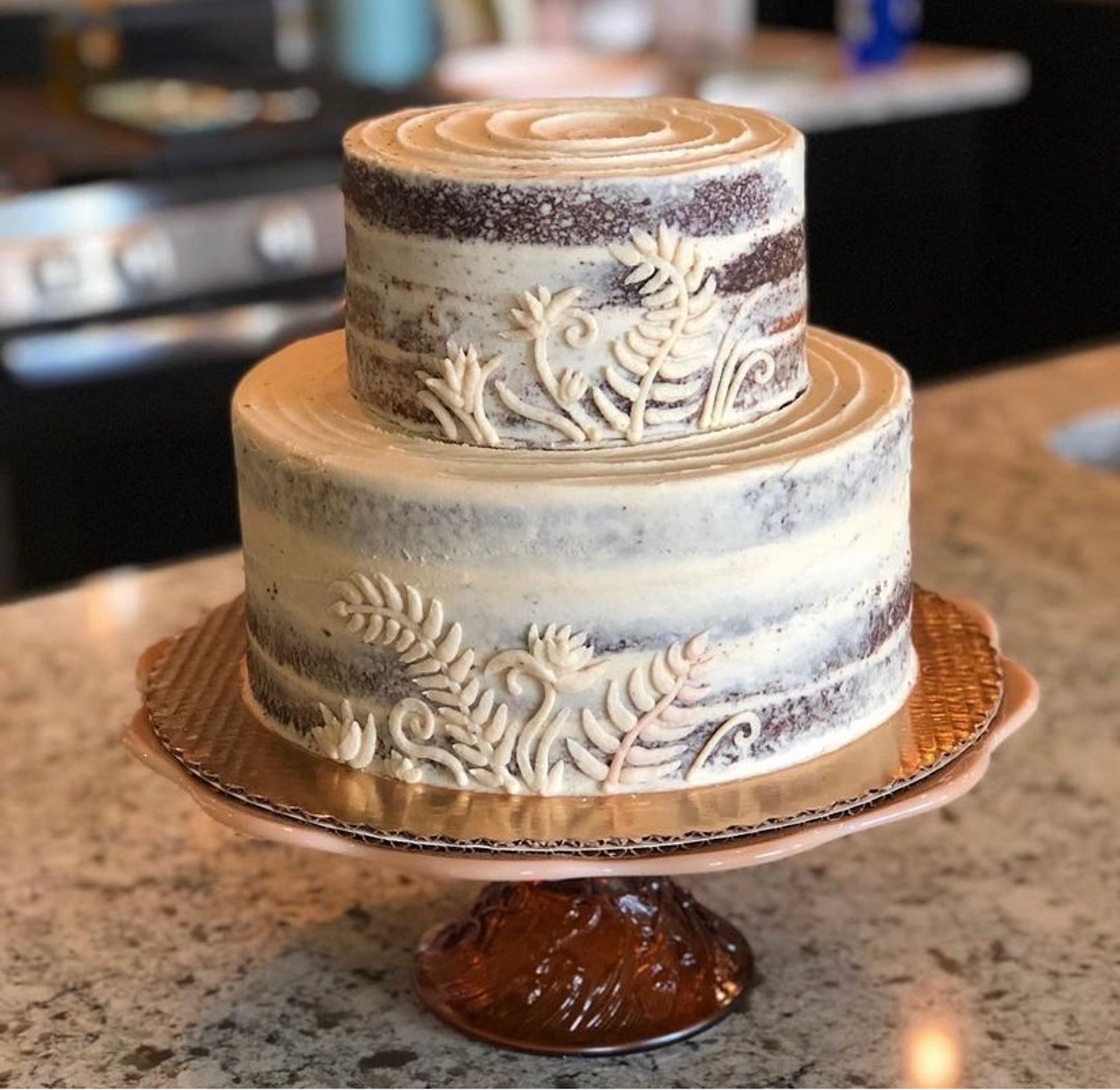 A two tiered wedding cake on a cake stand. Iced in a half naked style with layer silhouettes showing through, with botanical filigree layered over it in the same ivory colored icing.