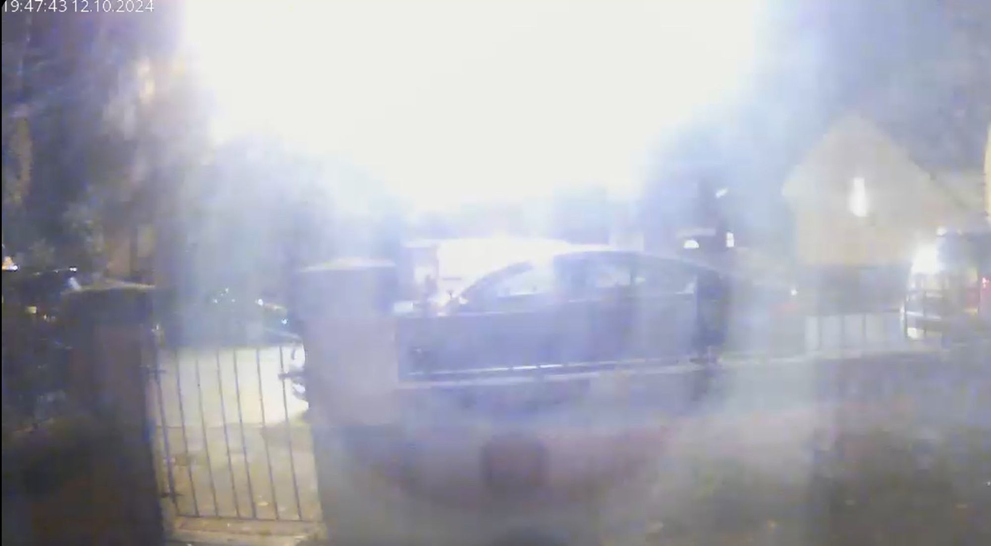 A low quality camera shot of a main road in England, from the view of a house on the street. Most of the frame is lit bright white from the rough center top where the sky it. 