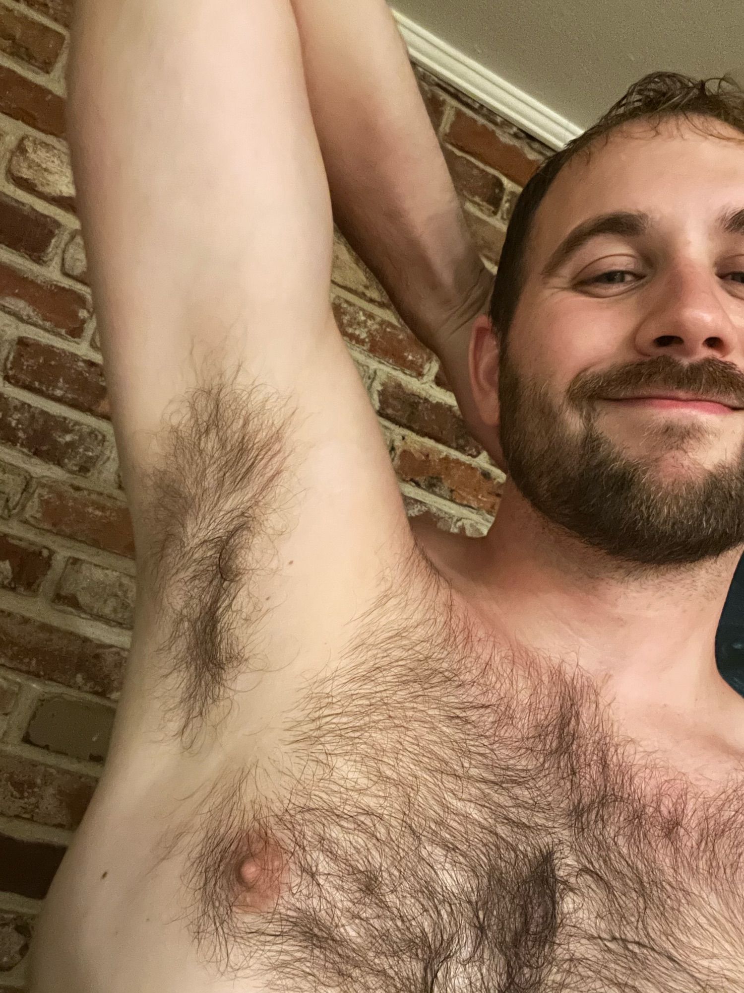 Rob is smiling against a brick wall. He's shirtless and has his right hand behind his head so as to show off his hairy armpit