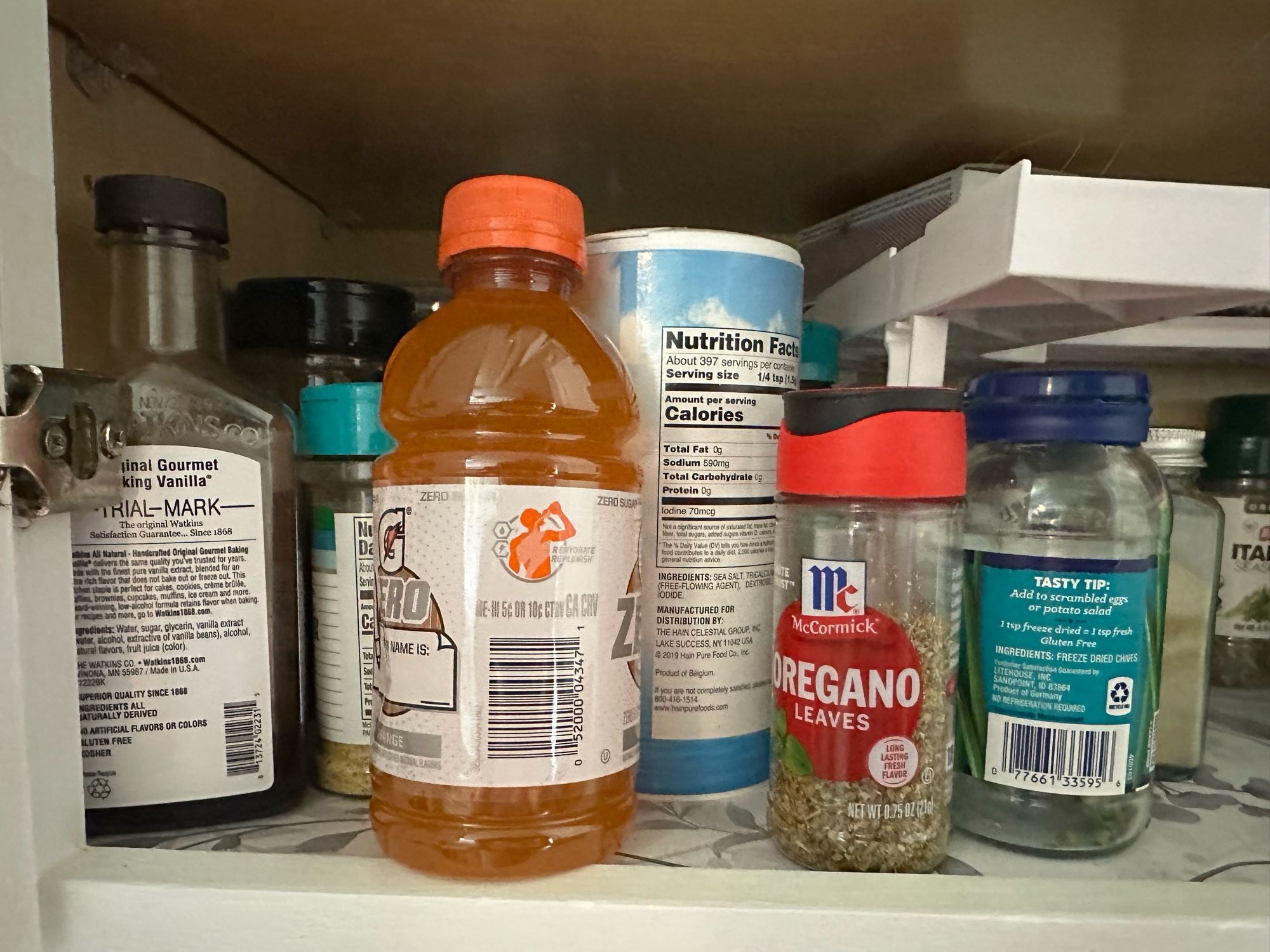 A bottle of orange Gatorade zero sitting among the spices in a spice cabinet.