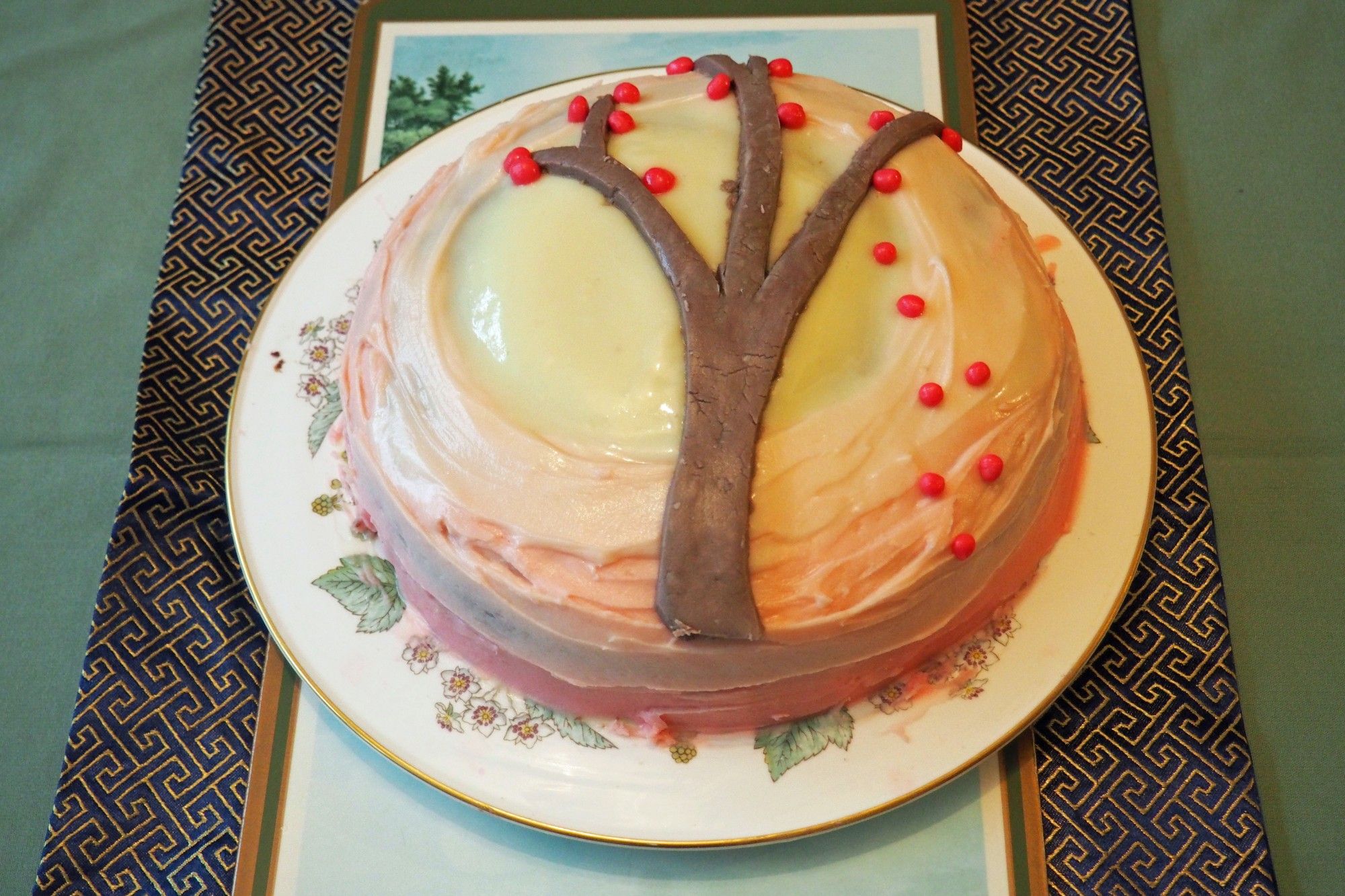 Autumn birthday cake on a plate. The cake is decorated in pastel shades of cream, pink and magenta, with a brown tree across the top, with red apples falling from the tree