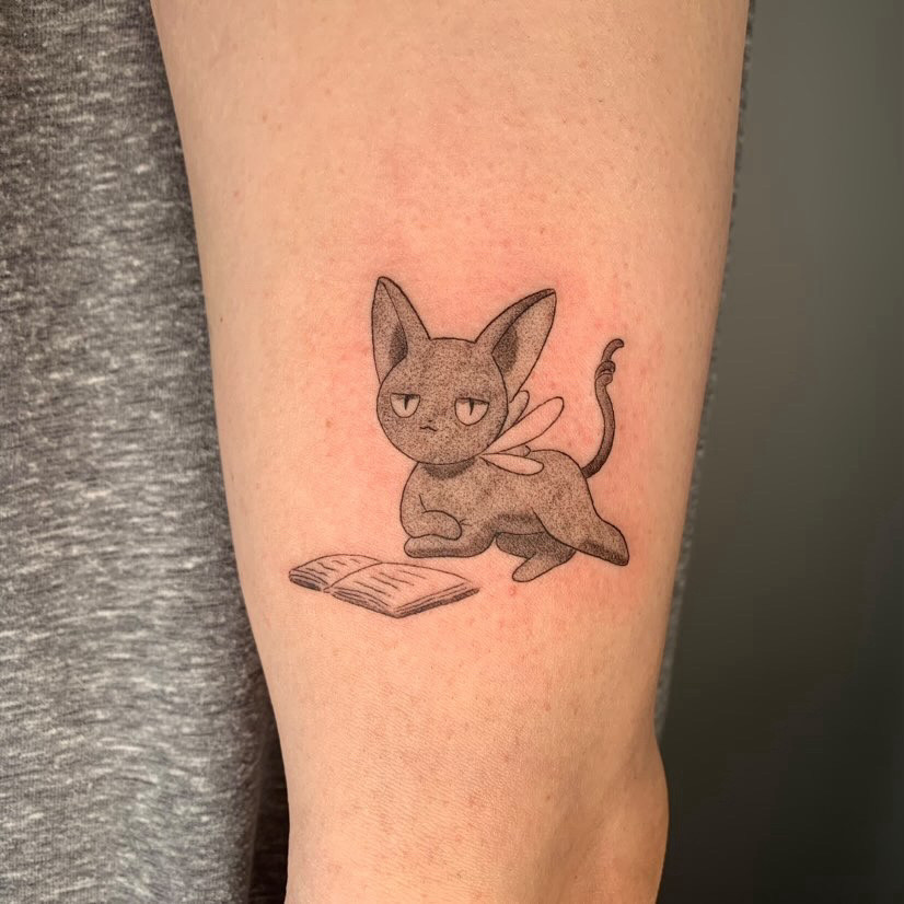 Foto de uma tatuagem do personagem Spinel Sun, deitado com as patas dianteiras cruzadas, em frente a um livro aberto. A arte é toda em escala de cinza, aplicada na lateral do bíceps de um braço de pele branca.