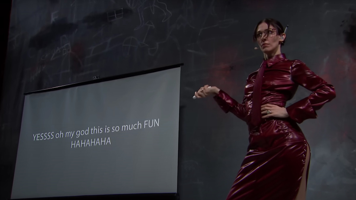 Singer-songwriter Caroline Polachek, wearing a skin-tight dark red leather piece that evokes stereotypical librarian outfits poses while presenting a slide-show. On the projector, we can read the words "YESSS oh my god this is so much FUN HAHAHAHA"