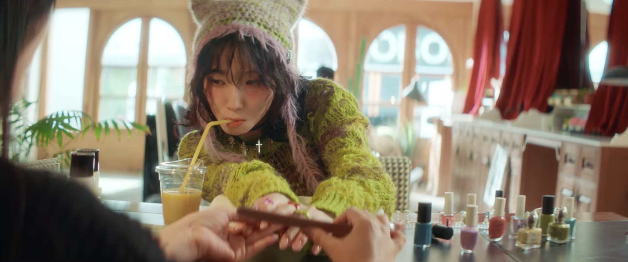 Kep1er’s Youngeun wearing a green sweater and a fuzzy hat getting her nails filed while she sips a drink from a straw.