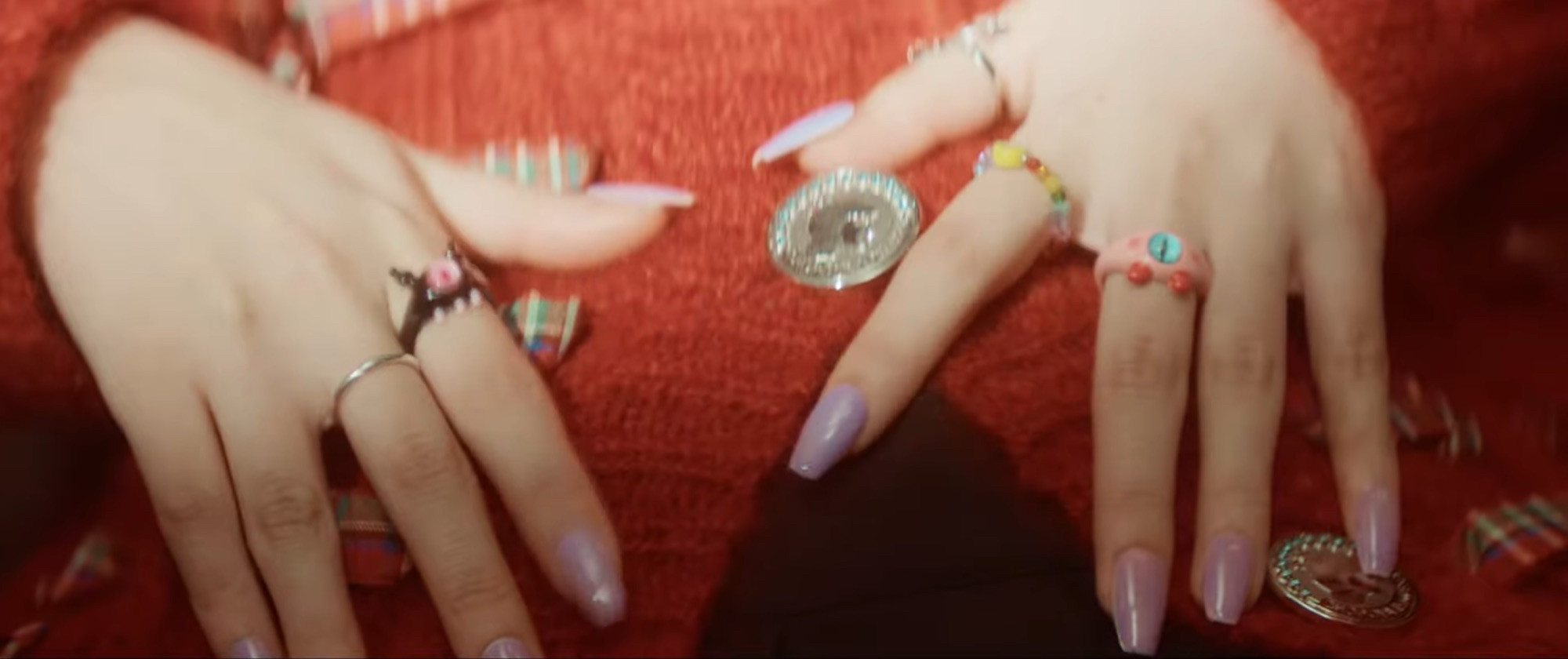 Kep1er’s Hikaru’s hands with multiple colorful rings on top of her red cardigan with large silver buttons.