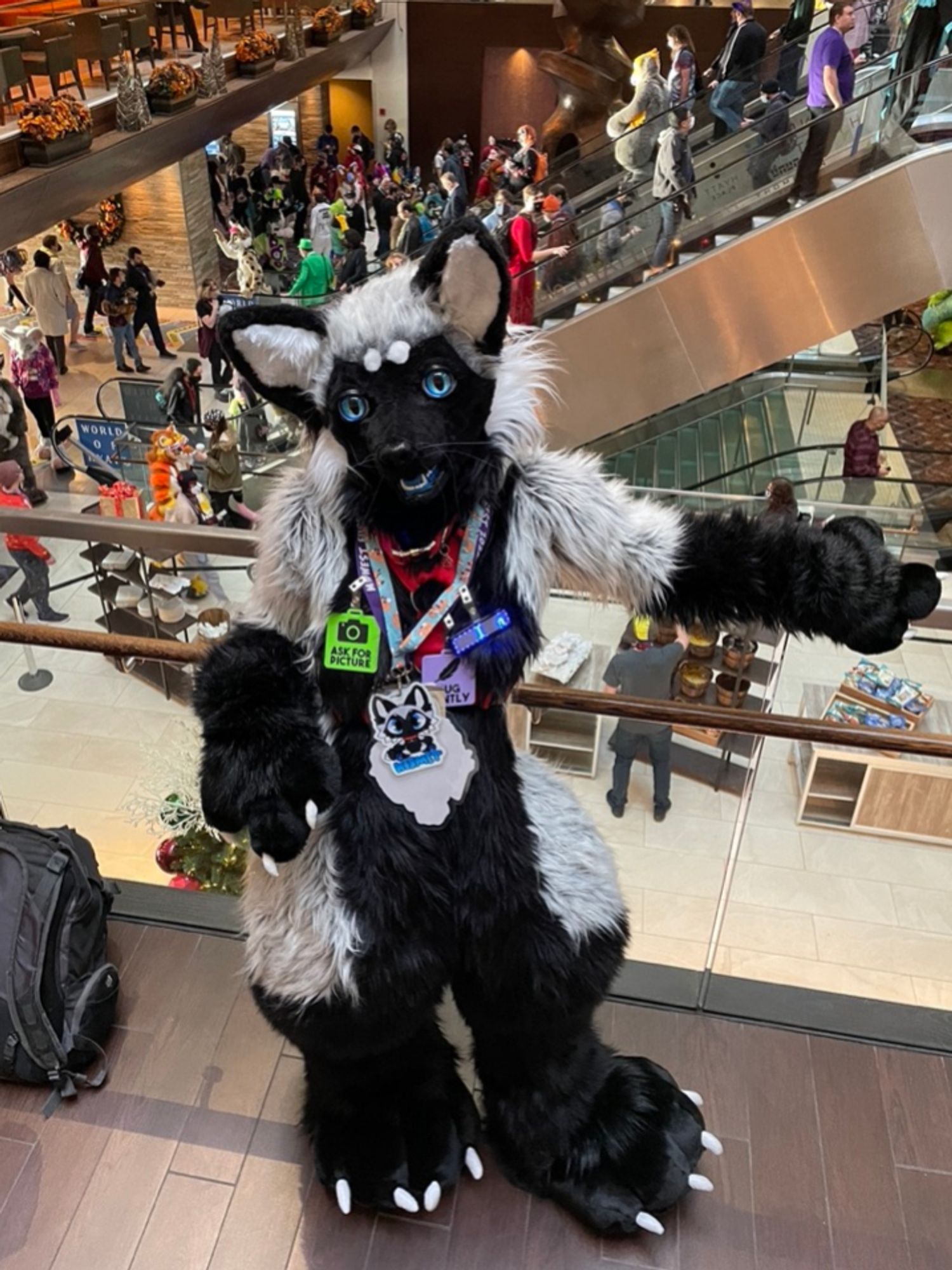 Silver fox fursuiter standing on a convention center balcony. There are people and other costumes of other species in the backgrounds. She looks uo at the camera and is covered in green, purple, black, and blue badge accessories. Her left arm rests on the wooden edge of the glass wall.