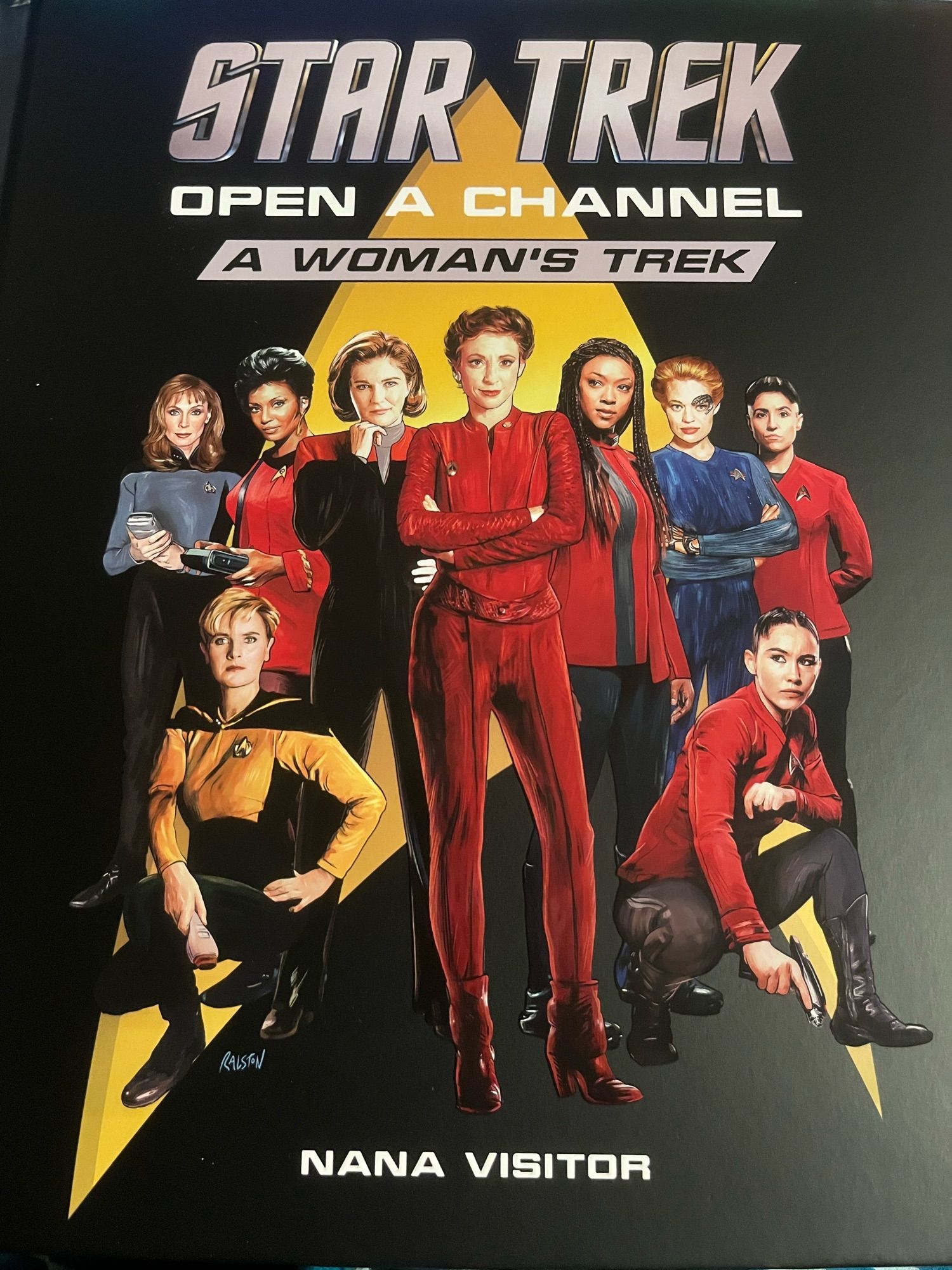 Cover of Star Trek Open a Channel A Woman’s Trek by Nana Visitor. Artwork shows a Starfleet logo in the background and in the foreground, Beverly Crusher (TNG era), Nyota Uhura (TOS era/Nichelle Nichols), Tasha Yar, Katherine Janeway, Kira Nerys, Michael Burnham, Seven of Nine (VOY era), La’an Noonien-Singh and Erica Ortegas