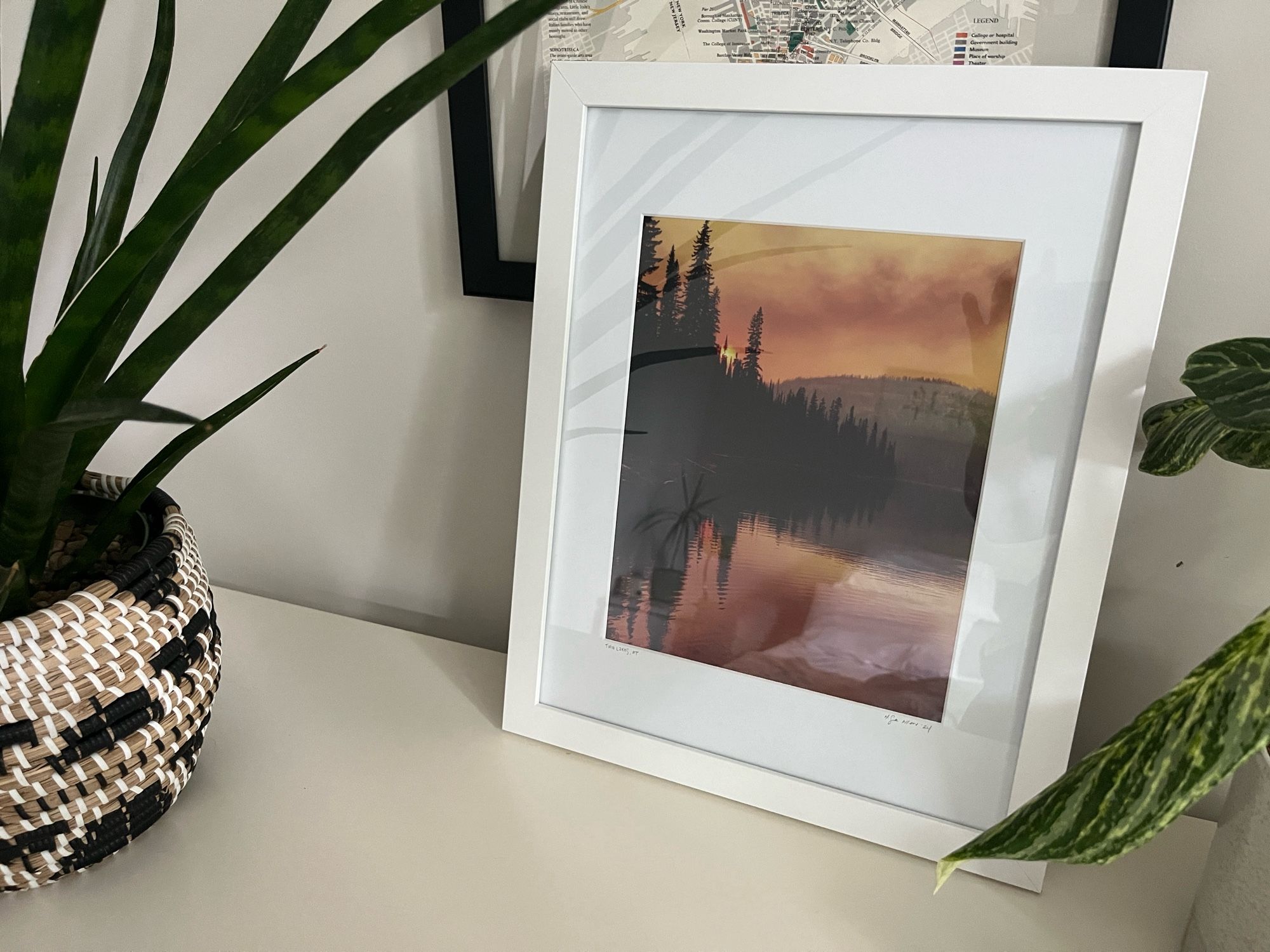 A framed print of a smoky sunset mountain lakeshore. The sky is orange and the pine trees on the left bank are dark silhouettes that reflect perfectly in the still water. The matte and frame are white. It’s sitting on a white dresser with a couple of plants, leaning up against a black-framed map.