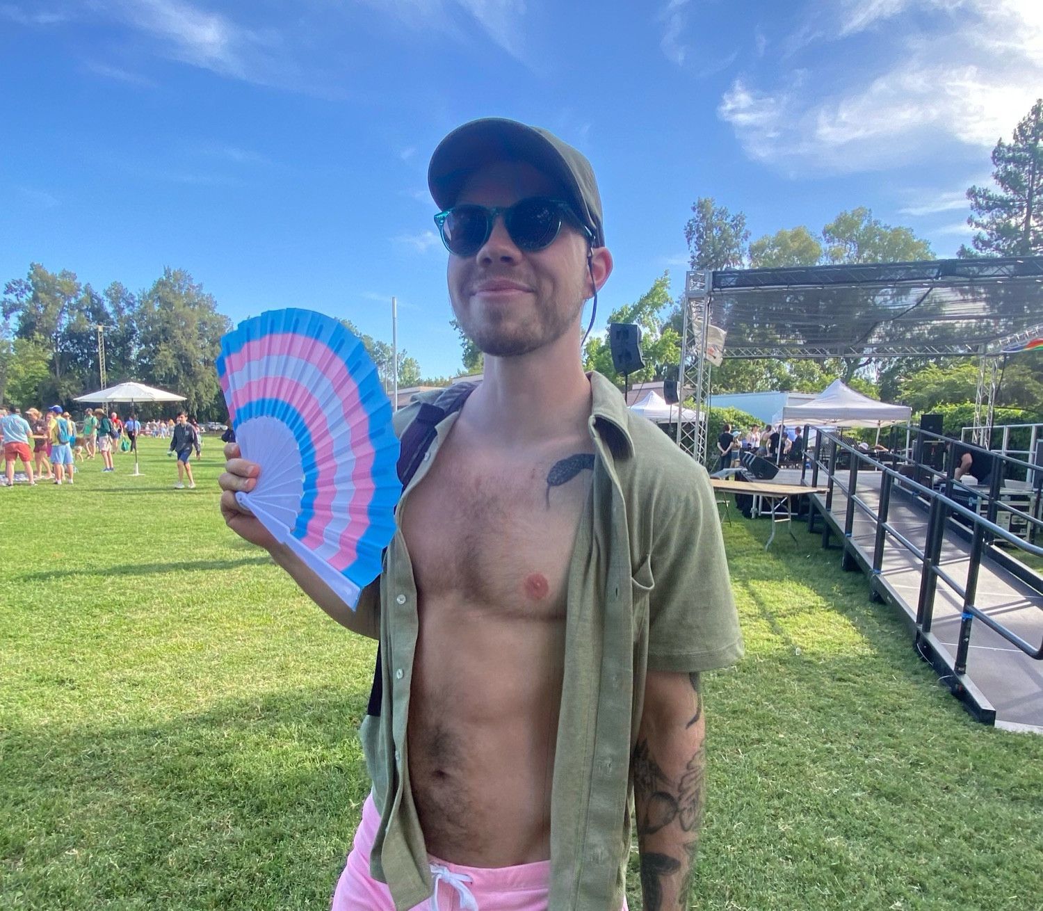 Shawn stands with button down shirt open and scars out on a grassy field with a stage and people behind. Hes holding a hand fan with a trans flag on it, wearing sunglasses and a baseball hat, and smiling