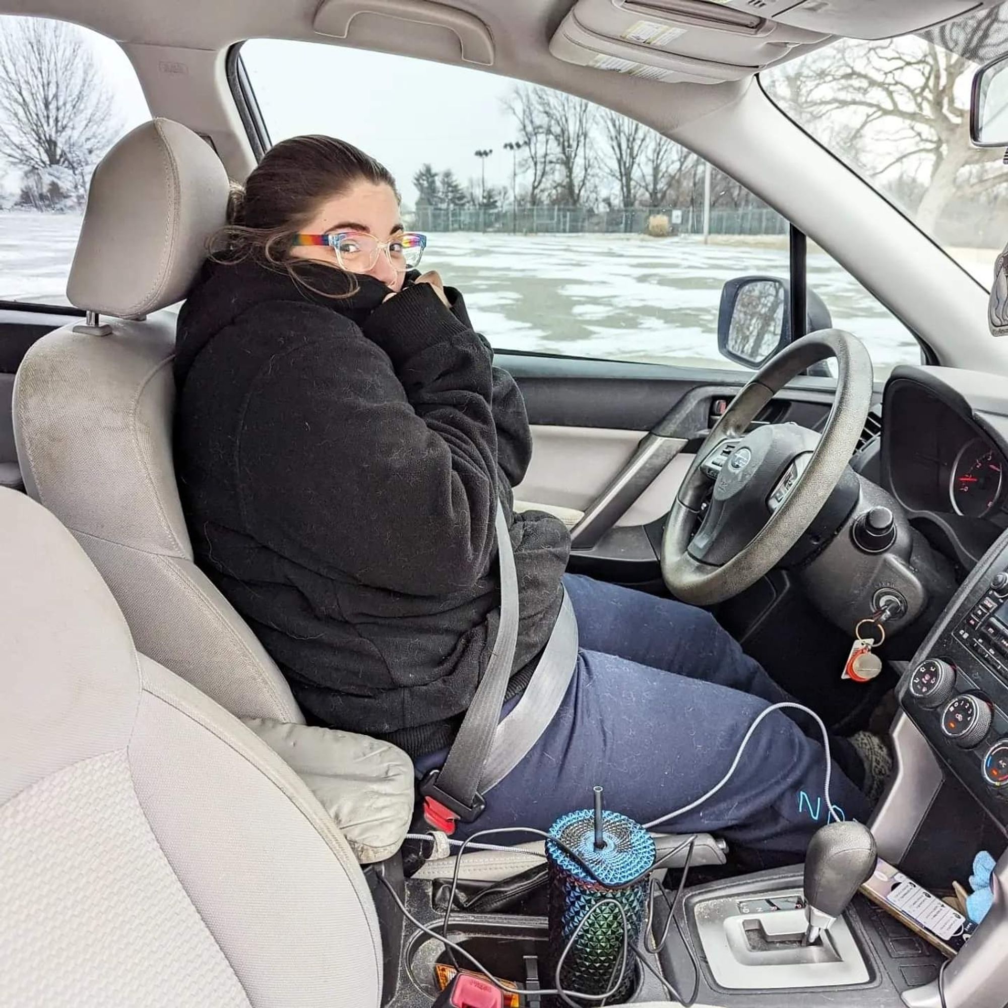 A white girl in a giant black hoodie looking cold and sitting behind the wheel of a car.