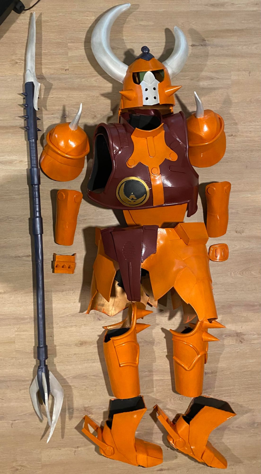 An orange and brown suit of EVA foam armor laying on a hardwood floor. A spiky weapon with one blade at one end and three on the other is beside it.