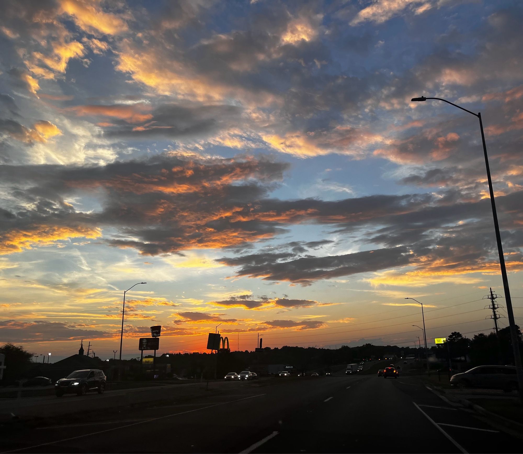 Photo of the sky at sundown, clouds are grey and orange. The road is dark and several vehicles have their headlights on.