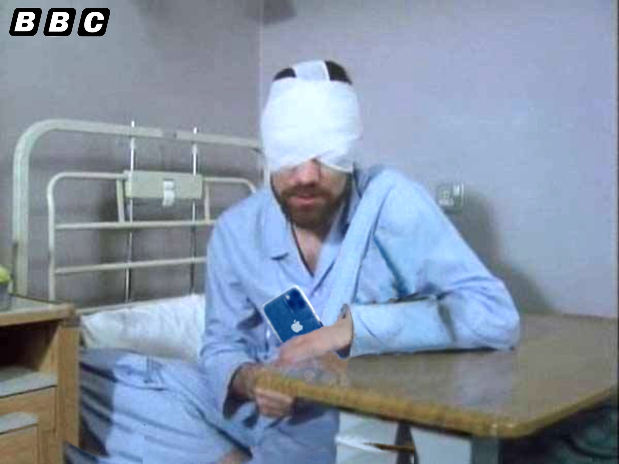 John Duttine BBC Day of the Triffids 1981 - a man with a beard, completely bandaged over eyes and upper head, sits up in a hospital bed leaning on a bed table dictating into a smartphone [originally a tape recorder]