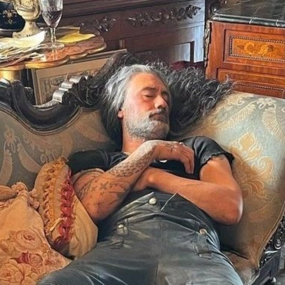A bts picture of Taika Waititi in costume as Ed reclining on a couch taking a nap.