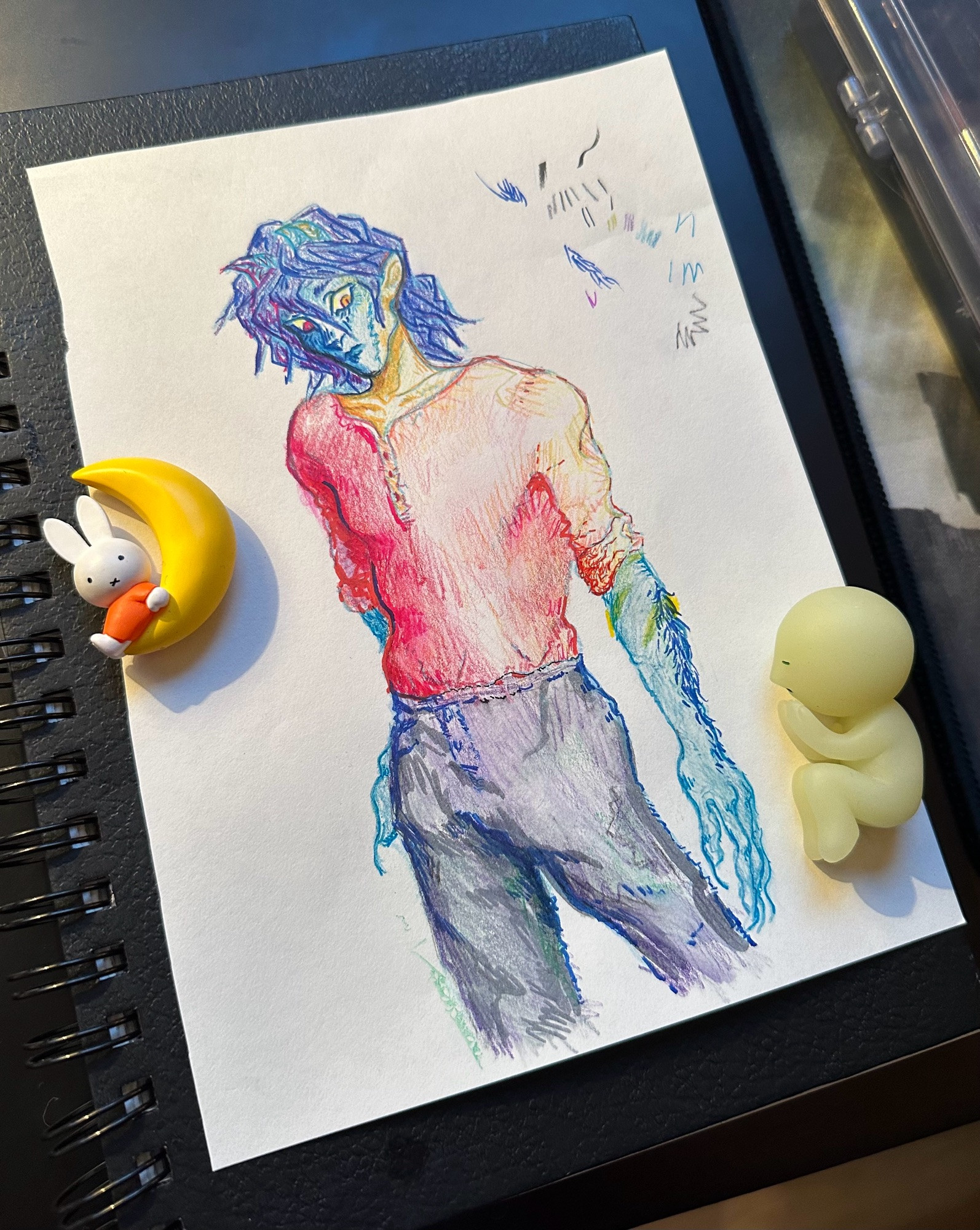 A photograph of a mixed media illustration. Atro, the artist’s original character, is drawn. He cat ear-shaped demon horns and stands looking moody. He wears a red shirt and gray pants. His skin is lemon-lime and light blue. His dark blue hair almost touches his shoulder. A tiny miffy ornament and a cartoon figure of a person sleeping sit on top of the paper. 