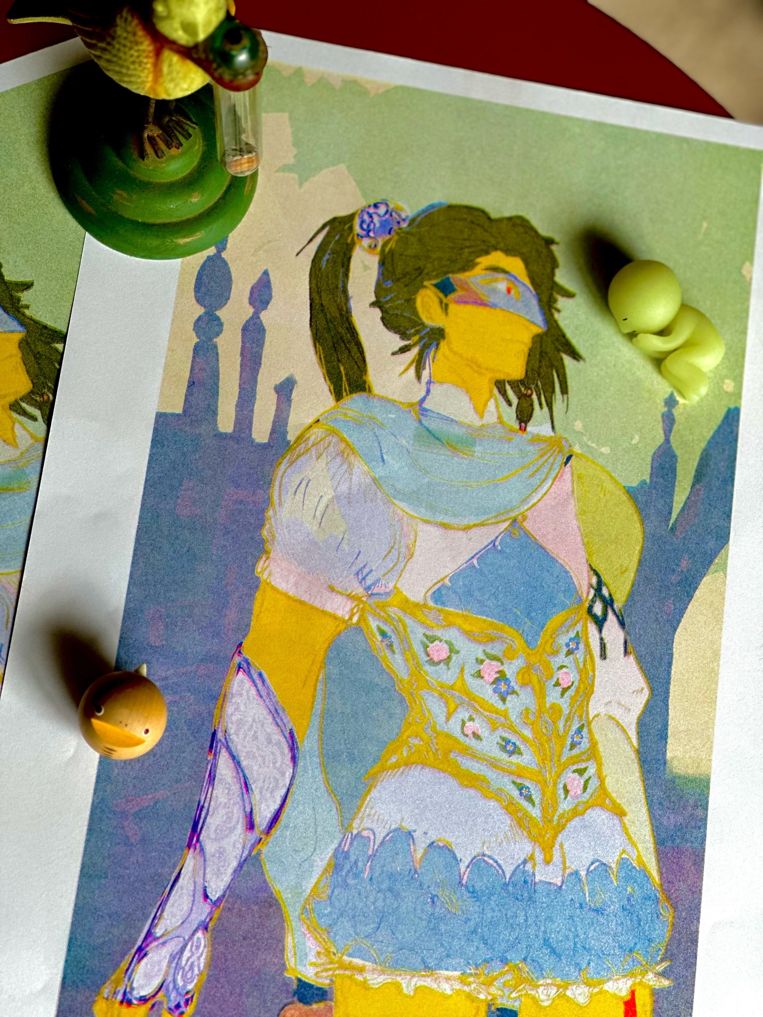 A photograph of a Riso print on a red table. The prints are fantasy illustrations of a young man with dark green hair and golden yellow skin. He wears an ornate pastel outfit comprised of blue pantaloons, a porcelain corset, and a shirt made of four diamonds meeting at his chest. He also wears a ceramic mask over his eyes and a porcelain gauntlet decorated with floral motifs on his right arm. Sitting on top of the prints are a small bird statuette, a tiny figure of a cartoon sleeping person, and a spherical snake.