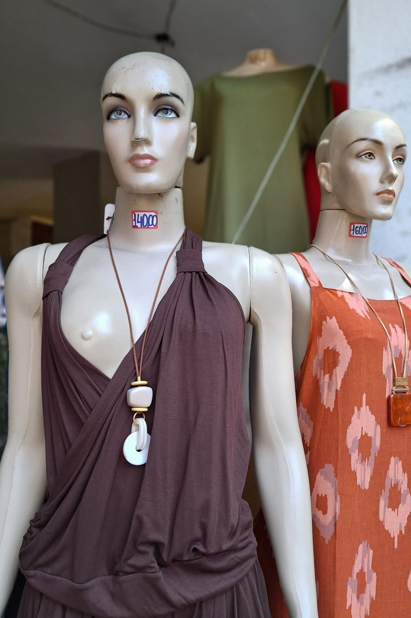 a foto mostra duas manequins de uma loja, uma em frente à outra. a de trás usa um vestido alaranjado; a da frente, um conjunto de calça e blusa sem mangas de cor marrom, e está com o seio direito aparecendo para fora da blusa.