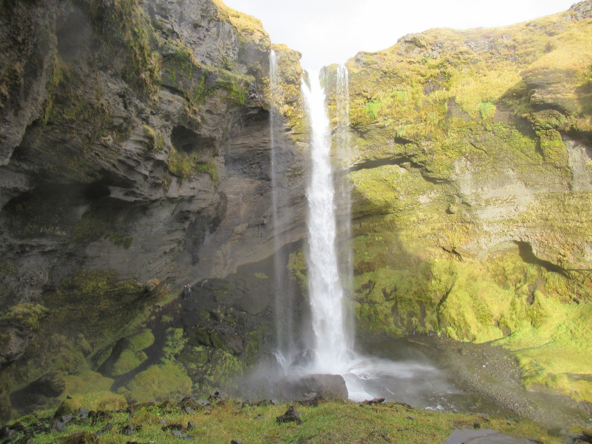 Kvernufoss, une cascade de 30 m dans un canyon verdoyant. 2 personnes se tiennent contre le rocher à gauche, pratiquement derrière la cascade. Islande (octobre 2018)