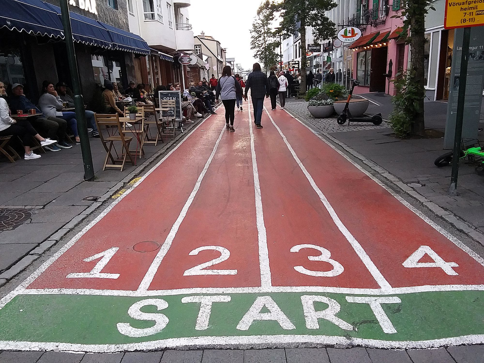 Piste de course sur la rue Laugavegur à Reykjavik, Islande. On voit le début du parcours (Start) et les 4 couloirs de course numérotés de 1 à 4. De chaque côté, on retrouve des commerces et une terrasse. (juillet 2022)