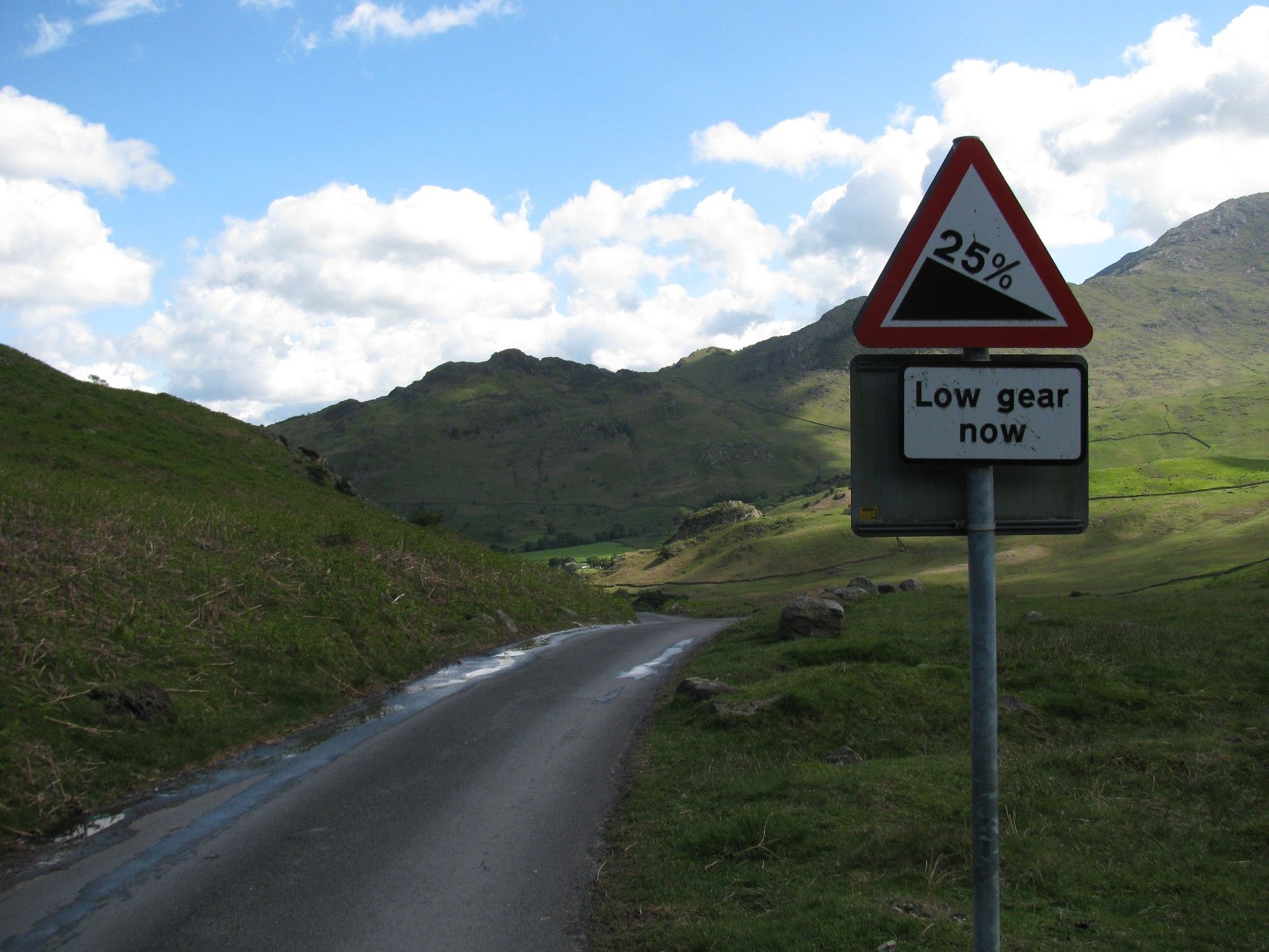 Une route étroite et sinueuse entre les collines. Le ciel est partiellement nuageux. Un panneau indique une descente à 25% et d'utiliser la première vitesse maintenant. Lake District, Angleterre (mai 2011)