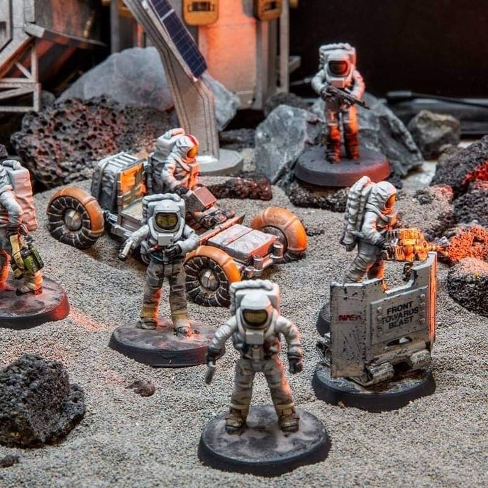 A promotional photo for the tabletop game Lunar by Black Site Studios. It shows several astronaut miniatures, a remote robot unit miniature, and an astronaut driving a buggy, all on a moon surface game board. 