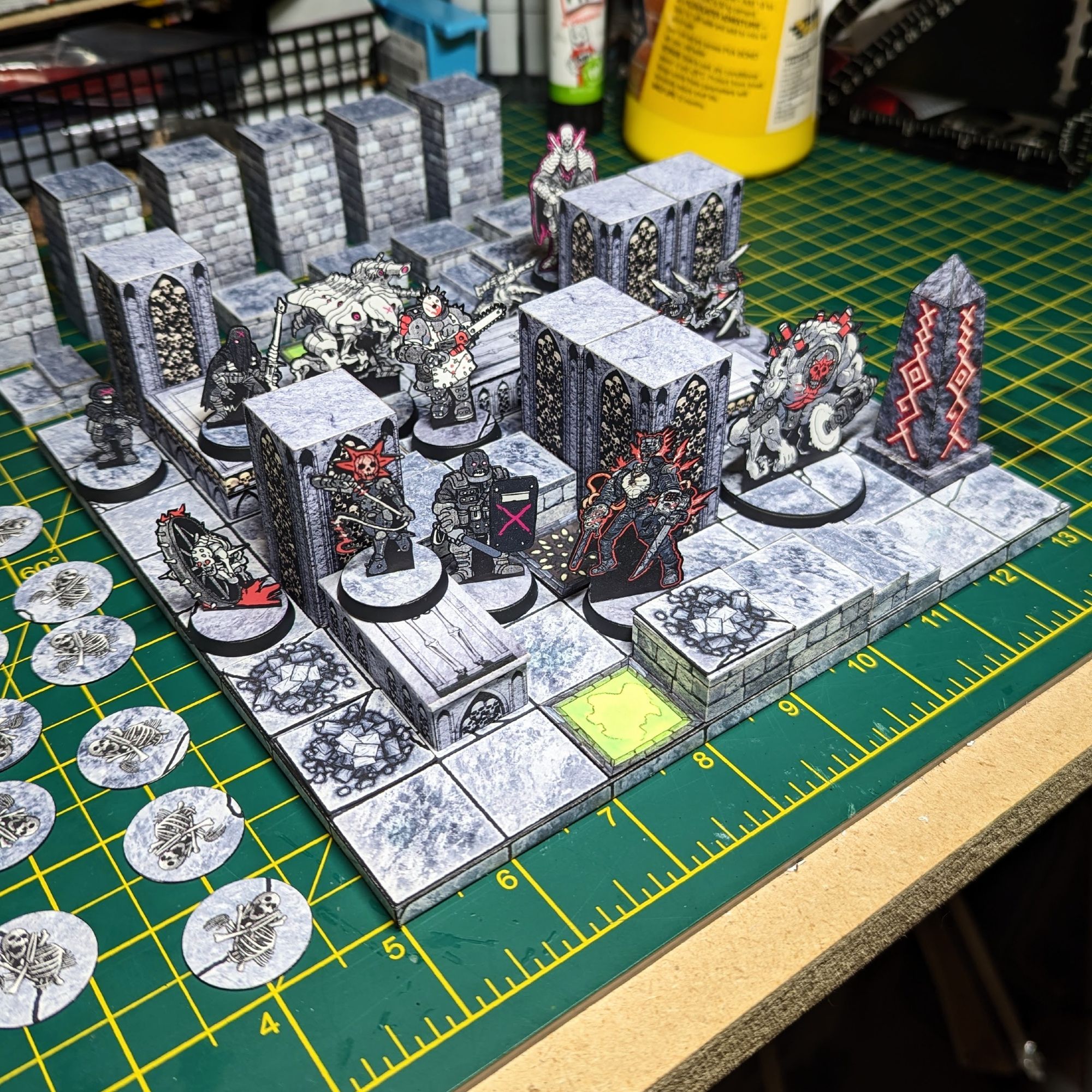 A photo of a 2.5D papercraft game board for Maleghast. A 8x8 grid of flagstones is filled with walls covered in skulls, tombs, raised areas with steps, and tokens representing rubble and pit traps. Populating the board are standee miniatures of two squads of various ghouls and monsters led by two Necromancers.