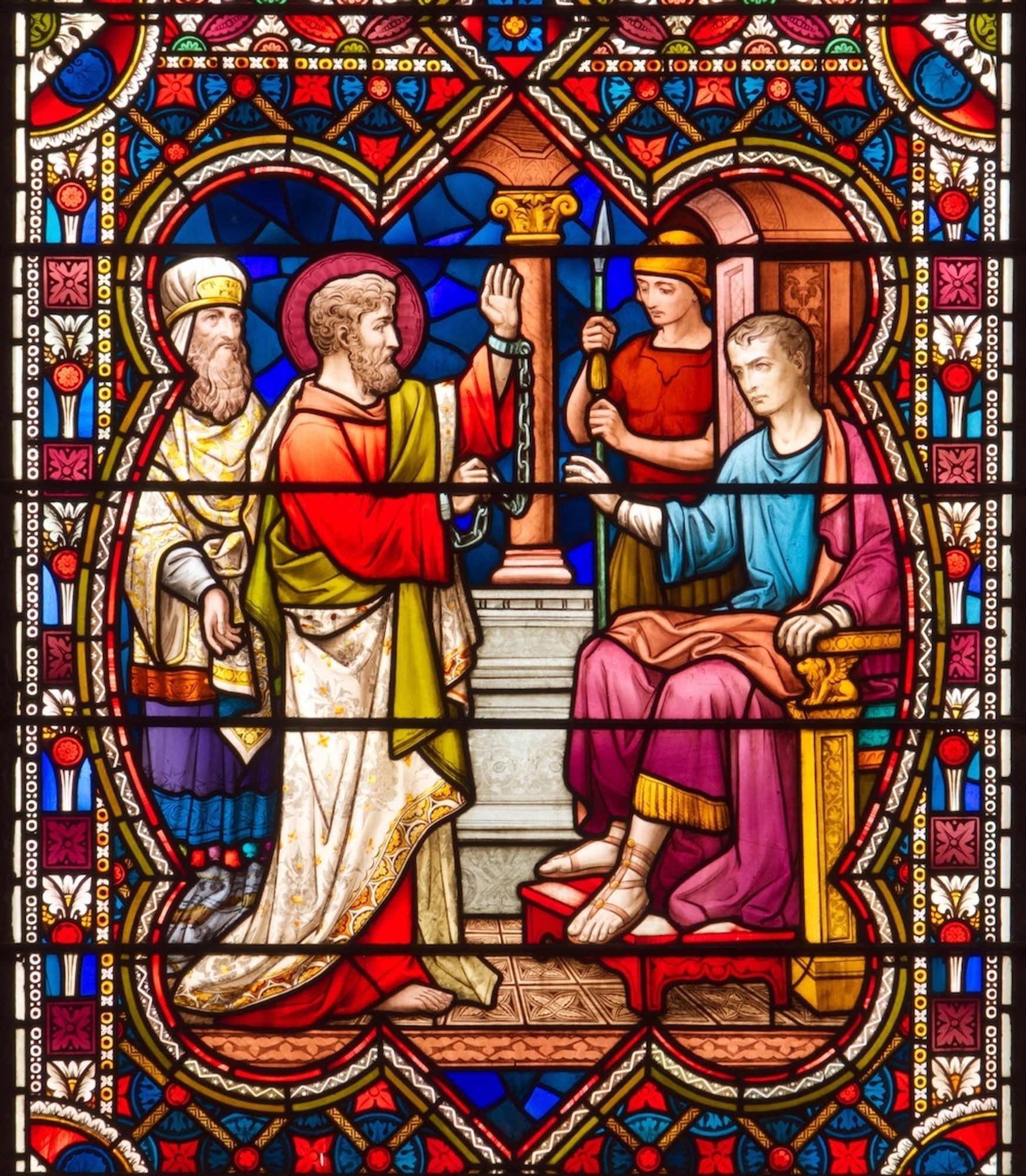 Stained glass panel of Saint Paul defending himself before Festus. In the foreground to the left Paul stands, facing the seated Festus on the left. Pauls arms are raised in rhetorical gesture, but his wrists are chained together. In the background behind Paul stands a figure in the dress of a Jewish priest, and behind Festus stands a Roman soldier with a spear.