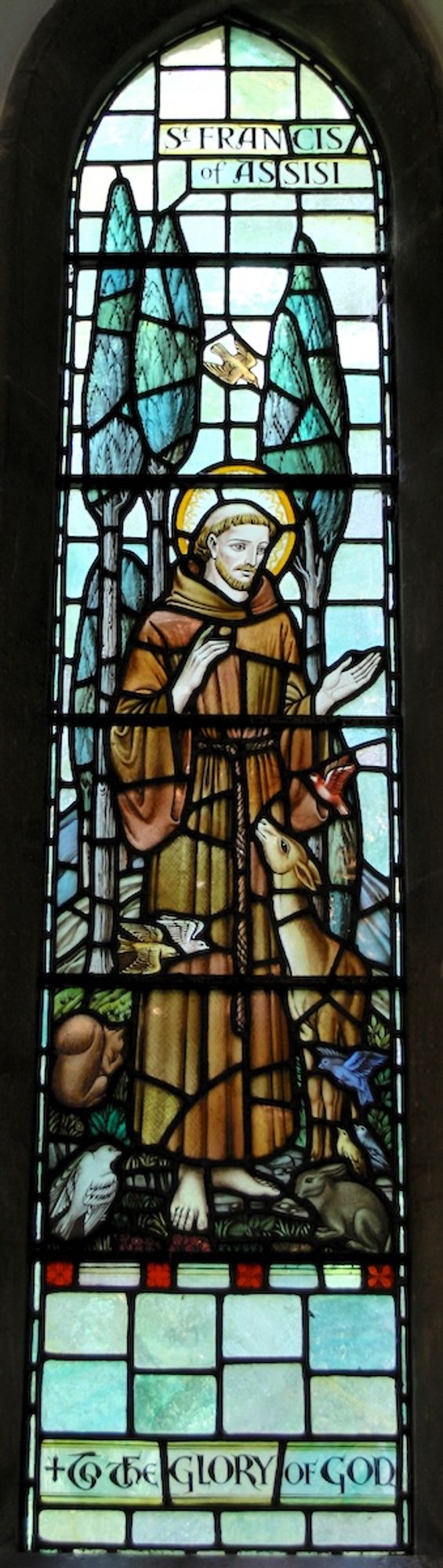 Staeind glass depicting Saint Francis of Assisi. The figure, clothes in brown monk's robes, stands surrounded by animals including a deer, a rabbit, and a squirrel, and several birds. There are trees in the background.