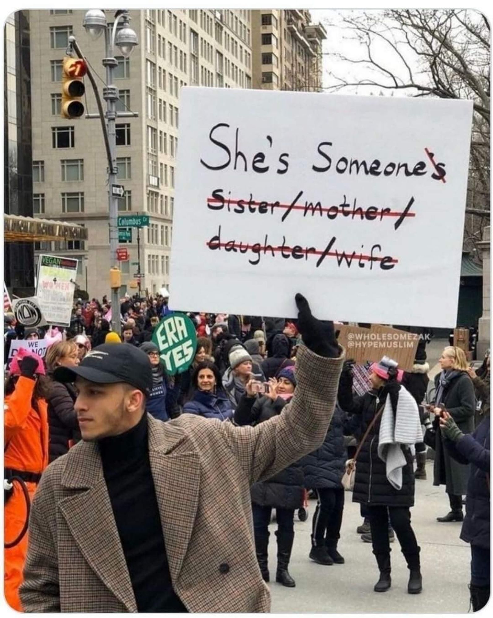 Man in a brown coat and black shirt and hat, holding a sign that says "She's someone's sister/mother/daughter/wife."
The list is crossed out so that the sign just says, "She's someone."
There's a crowd of people behind the man and one is holding a sign that says ERA YES.