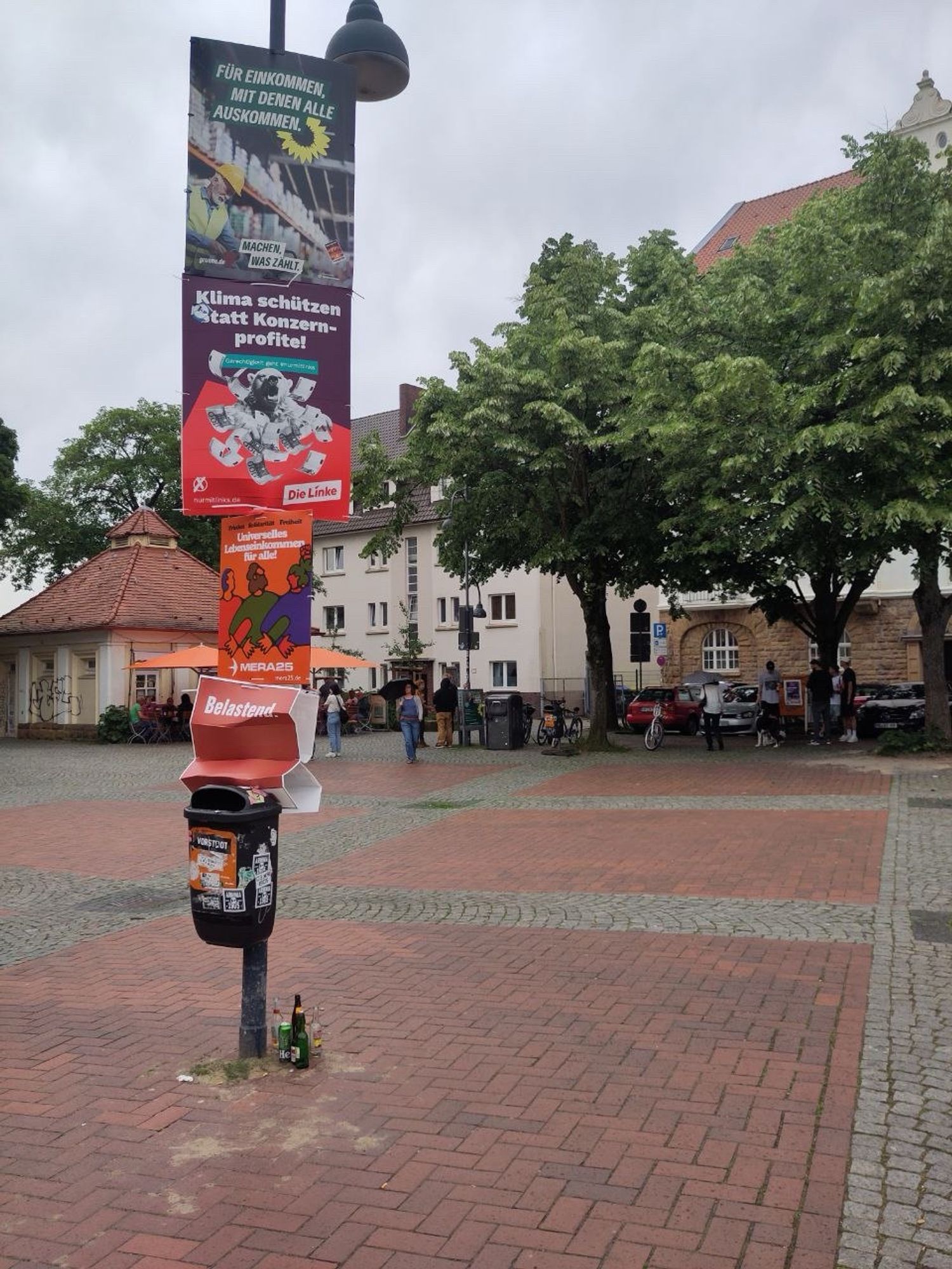 Ein Laternenmast mit vier Wahlplakaten und einem angehängten Mülleimer. Das untere Plakat ist von Die PARTEI und ist unter der Last der anderen drei zerquetscht. Es trägt die Aufschrift „Belastend“..