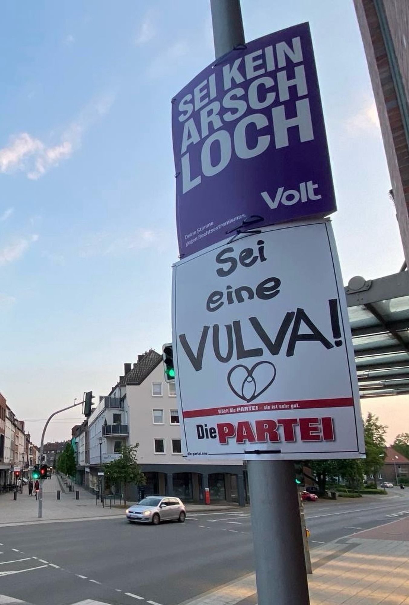 Ein lila Plakat mit der Aufschrift „Sei kein Arschloch“. Darunter ein Plakat von Die PARTEI mit dem Hinweis „Sei eine Vulva!“