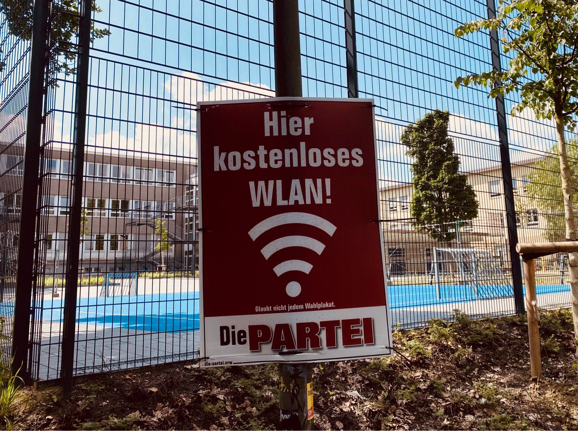 Ein Plakat mit WLAN-Symbol und der Aufschrift „Hier kostenloses WLAN!“, darunter sehr klein die Zeile „Glaubt nicht jedem Wahlplakat.“ Das Plakat hängt unmittelbar vorm Zaun eines Schulhofs mit Fußballfeld und kleinen Kindertoren.