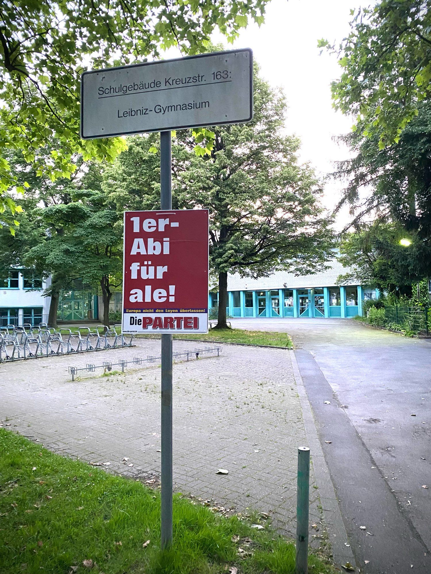 Ein PARTEI-Plakat mit der Aufschrift „1er-Abi für ale!“ vor einem Gymnasium