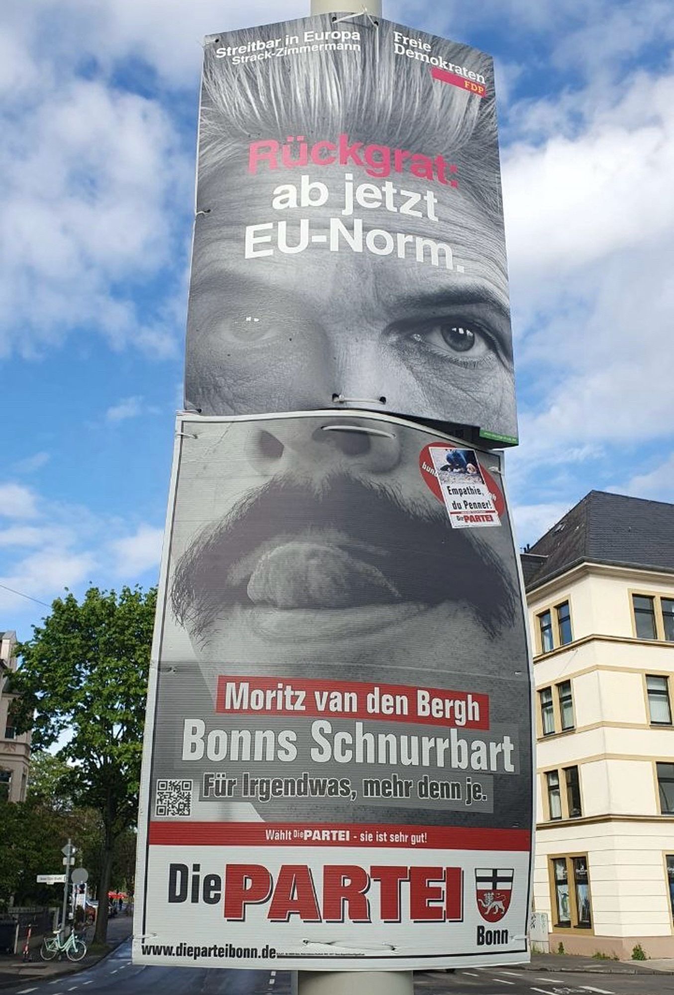 Ein graues Plakat, von dem die kalten Augen Strack-Rheinmetalls starren. Darunter ein graues Plakat von Die PARTEI Bonn. Motiv: Ein Mund, leicht geöffnet. Die herausgestreckte Zunge biegt sich hoch zum Pornobalken. Darüber der Blick in zwei beeindruckende Nasenlöcher.  Aufschrift: „Moritz van den Bergh Bonns Schnurrbart Für Irgendwas, mehr denn je. Wahlt dePARTEI - sie ist sehr gut!“