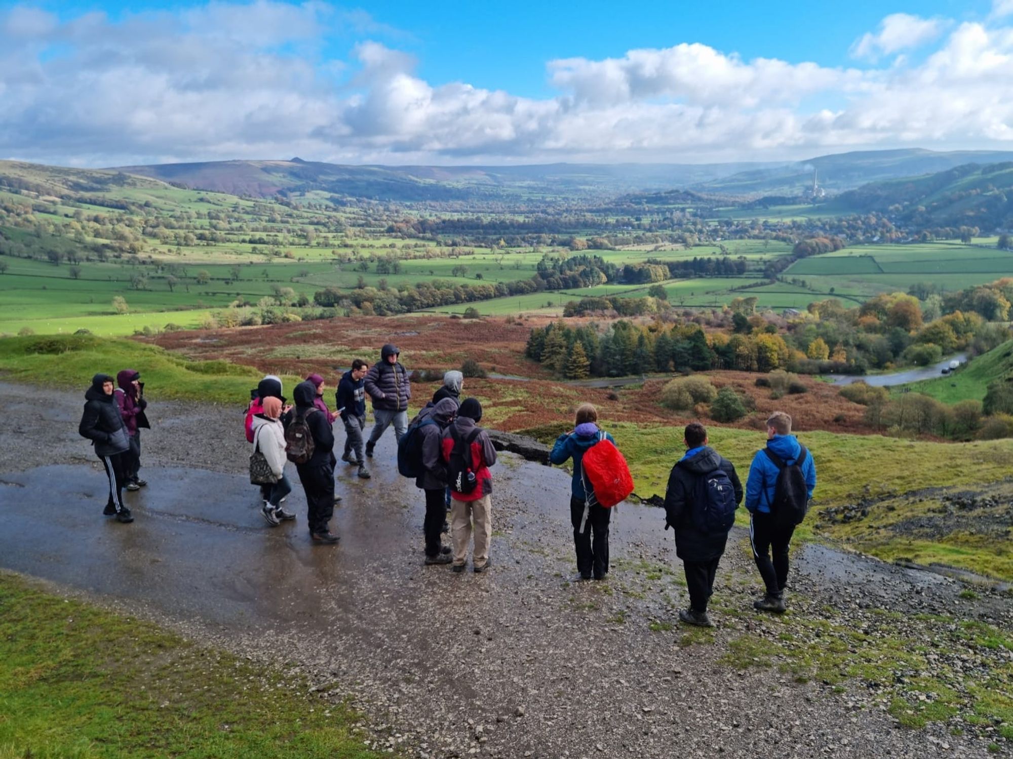 Students stand on a tarmac road that has cracked, slipped and fallen downhill. Hummocky landforms in middle ground covered in bracken form oart of the same mass movement event. Flat bottom valley with agriculture and chimney of a cement factory in distance (Hope Valley, UK)