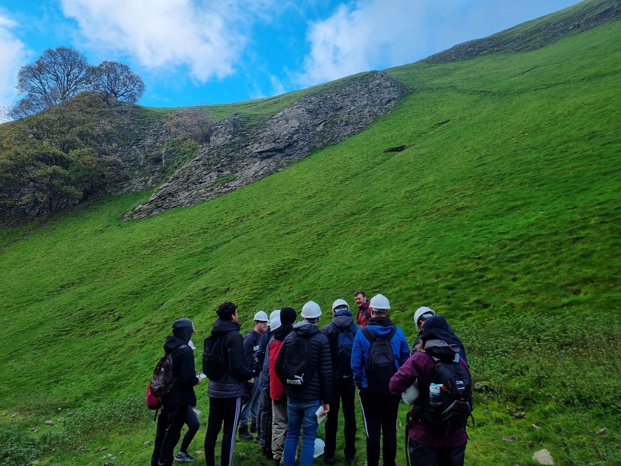 Group of students stand in front of a grassy slope and grey rock cliff faces in a limestone valley, Cavedale in Castelton, Peak District