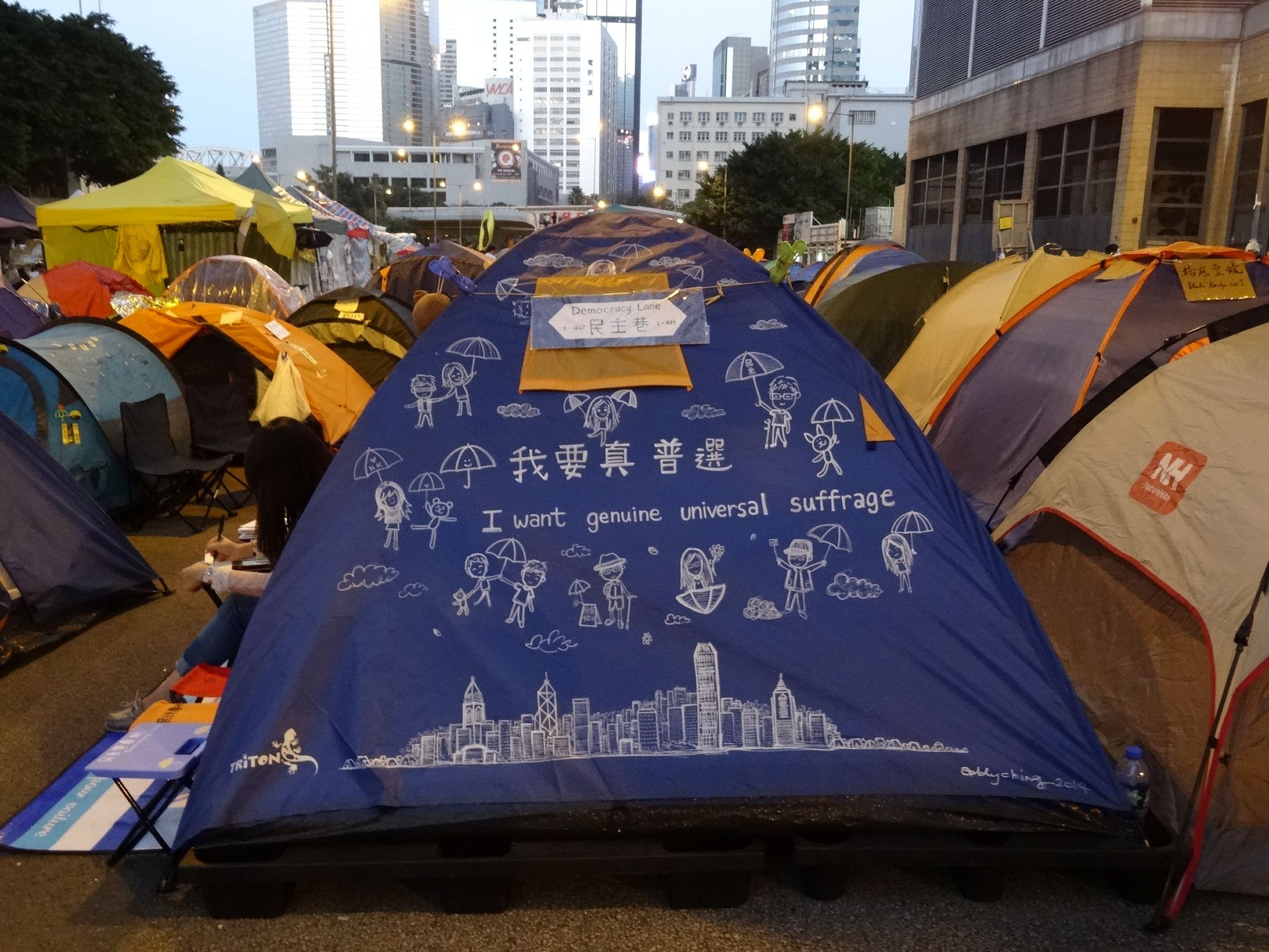 Tent on Harcourt Road, Admiralty, with cartoon illustrations of figures holding up umbrellas and also the city, and the words "I want universal suffrage" on it.