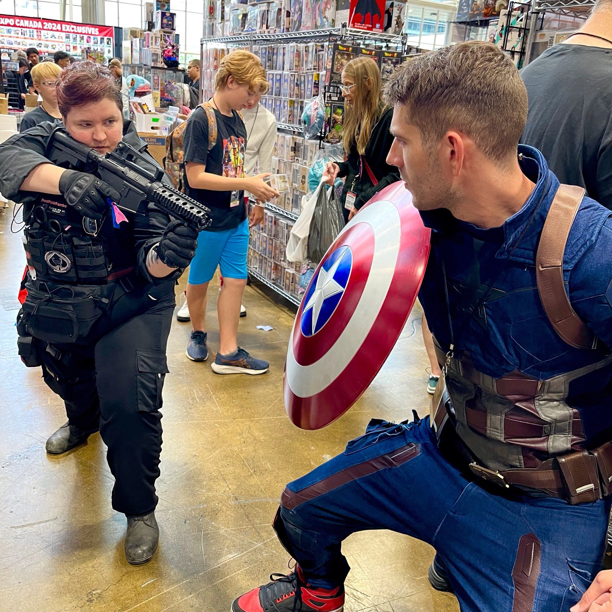 A woman in CALL OF DUTY armour aims her rifle at a shield carried by a Captain America cosplayer